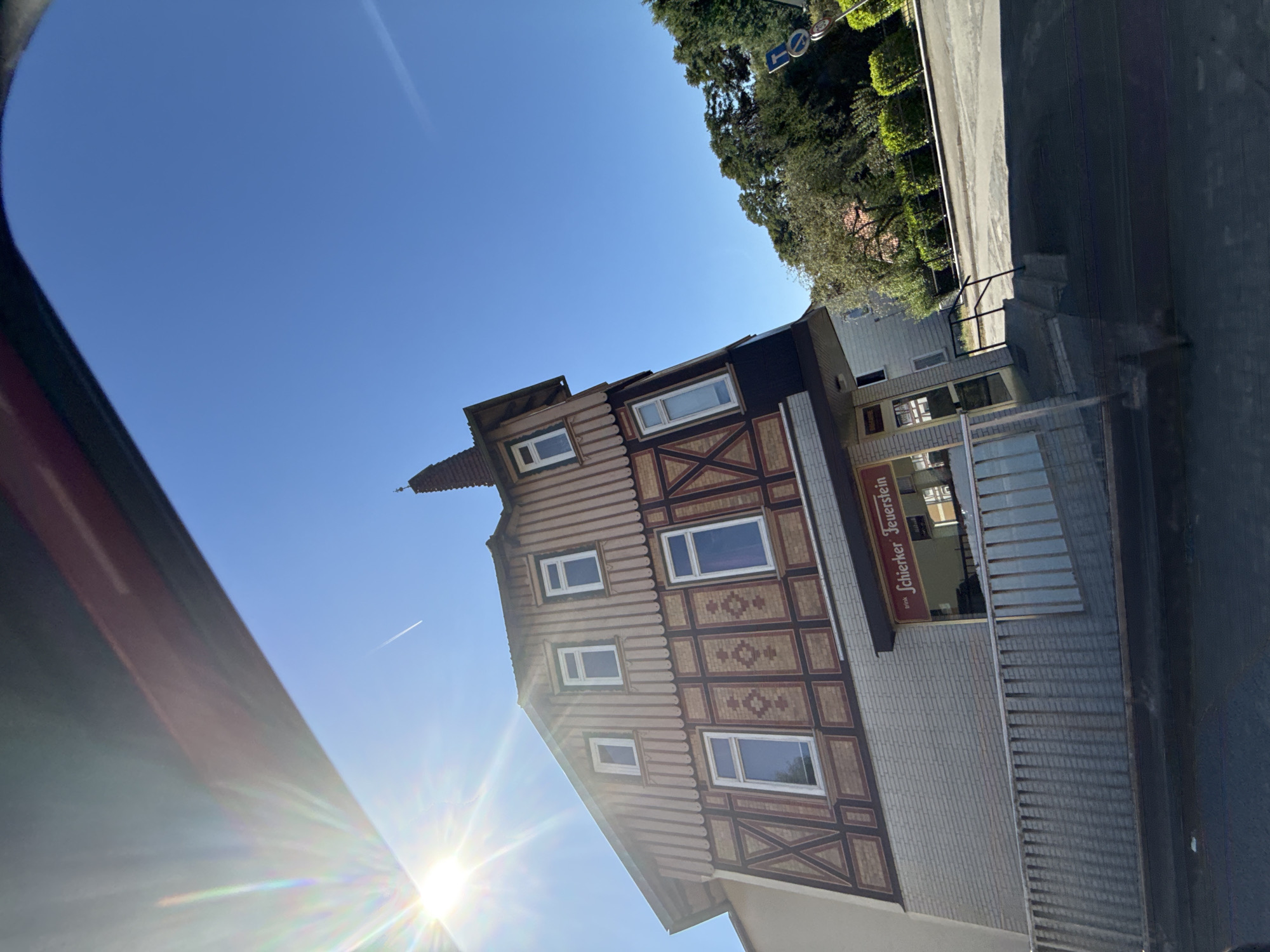 The image is a sunlit, slightly tilted, eye-level exterior shot of a building with a distinctive half-timbered upper facade and a brick lower section. The building is located in Bad Lauterberg im Harz, Germany. A sign above the entrance reads "Trink Schierker Feuerstein". The sky is clear blue with a faint contrail visible. Sunlight glares intensely from the upper left corner, creating lens flare. Green trees and bushes are visible in the background to the right of the building. A street and sidewalk area are in the foreground, with a metal railing in front of the building's lower level. No people are clearly visible.