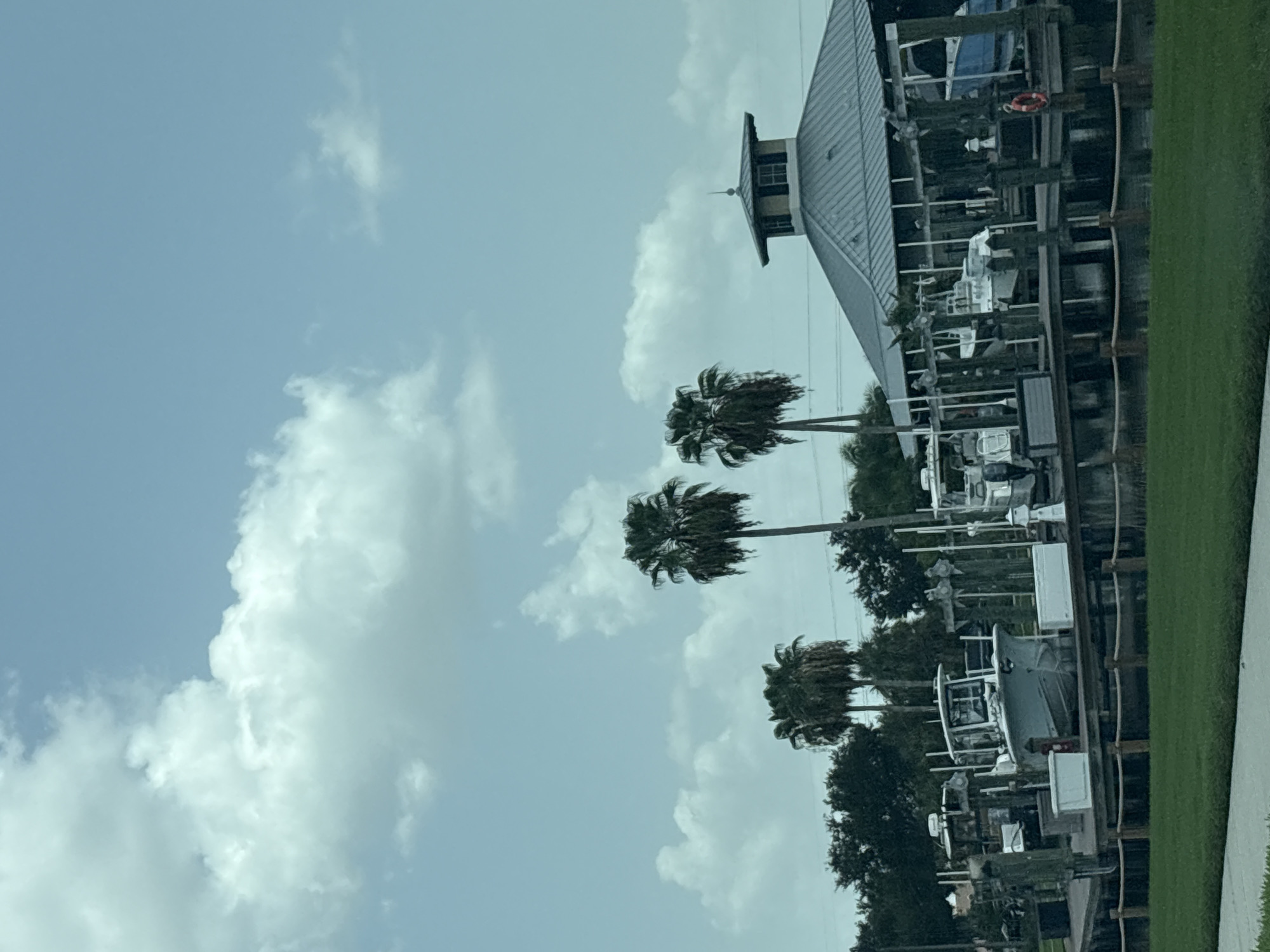 The image shows a marina on a cloudy day in Bradenton, Florida, United States of America. Several boats are docked at the marina, including a white center console boat in the foreground. A large, modern building with a distinctive roofline is visible in the background. Palm trees are scattered throughout the scene, and a wooden fence with rope accents lines the edge of the marina. The water is calm, reflecting the overcast sky. There are no people visible in the image.