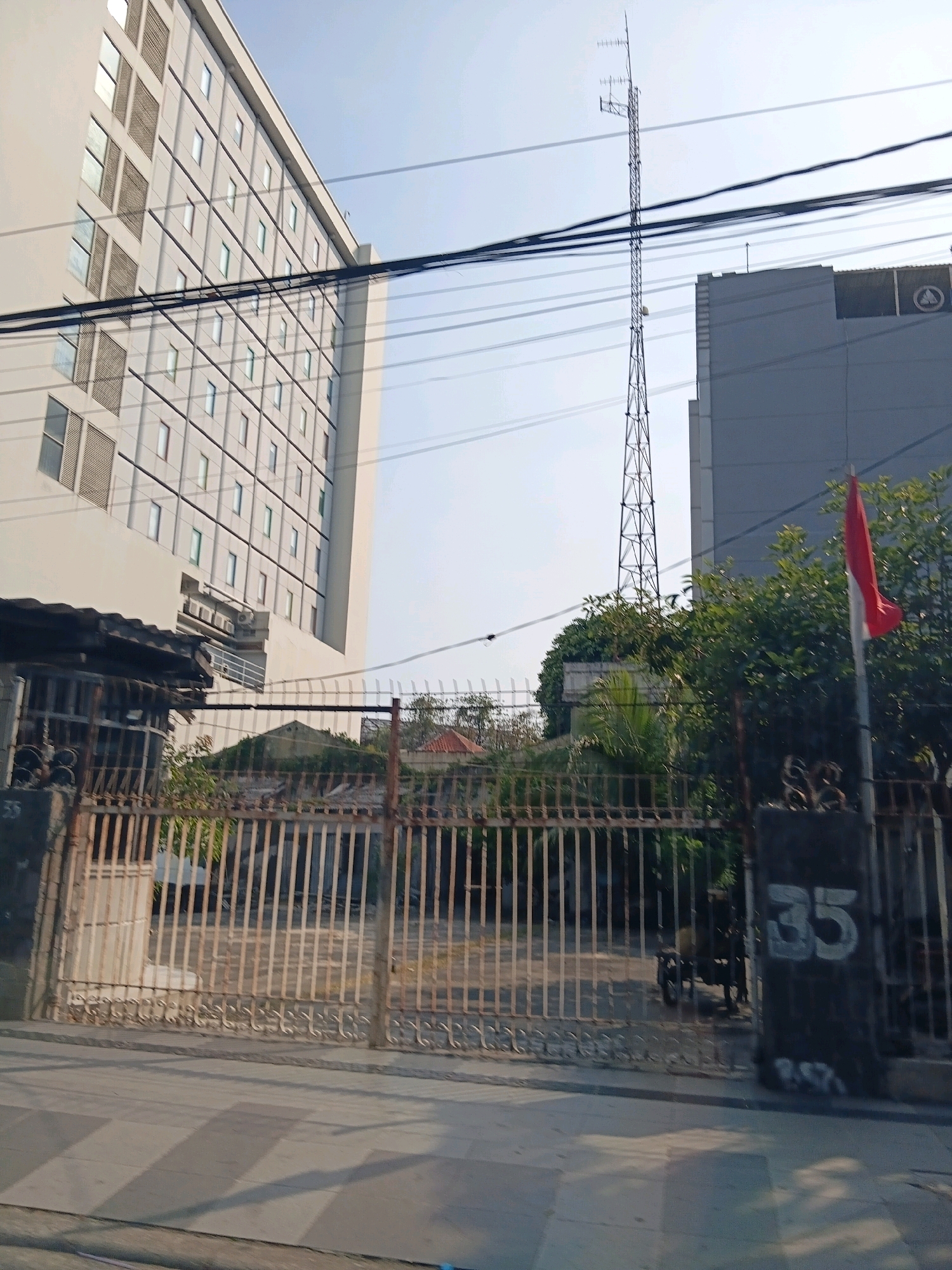 The image depicts a street scene in Surabaya, Indonesia, on a clear day. In the foreground, a rusty metal gate with decorative elements and the number "35" is visible. Beyond the gate, there is a glimpse of an overgrown lot with a small building. To the left, a tall, modern building with numerous rectangular windows and gray vertical and horizontal paneling dominates the frame. To the right, another building, also gray, is partially visible. A tall, slender transmission tower stands prominently in the background. A red and white flag, likely the Indonesian flag, is flying from a pole to the right of the transmission tower, partially obscured by trees. Power lines crisscross the sky. The street in front of the gate appears to be paved, with shadows cast across it, suggesting a sunny condition.