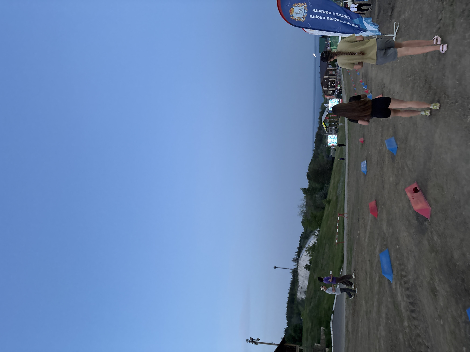 Captured in Khvalynsk, Russia, the photograph shows an outdoor race with participants navigating a course marked by red and blue flags. Two women jog near the top of the frame. One wears a yellow shirt with shorts, while the other is clad in black. Further down the path, a pair run side-by-side, one in purple. A large sign stands at the top corner of the image, and the sky is blue in the background.