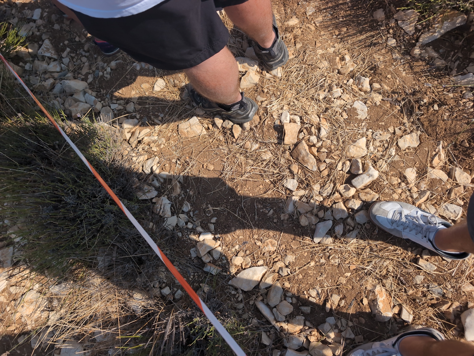 An overhead shot captures a close-up of a rocky, dry, outdoor path in Saint-Trinit, France. A white and orange striped ribbon is stretched across the ground, marking a boundary or course. The path is littered with small, light-colored rocks and dry, brittle grass. Shadows of people are cast across the scene, suggesting they are standing nearby, possibly participating in an outdoor event. The focus is on the feet and lower legs of at least three individuals, clad in athletic shoes and dark shorts. One person wears black shorts and dark, rugged trail running shoes. Another person's legs are visible, wearing light-colored athletic shoes and dark shorts. A third pair of legs, also wearing light-colored athletic shoes and dark shorts, is also visible. The overall impression is one of an outdoor race or trail event in a dry, possibly hot, environment.