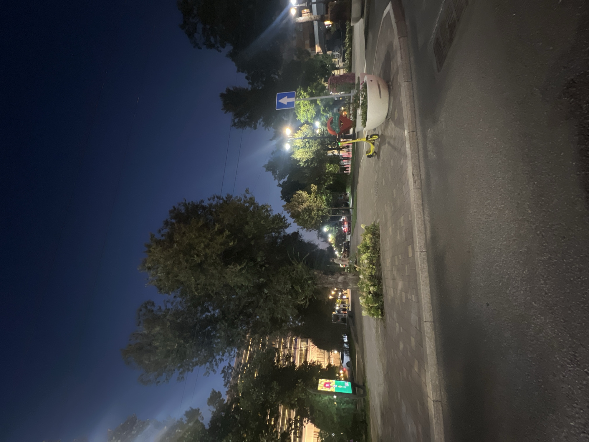 The image depicts a street scene at dusk in Almaty, Kazakhstan. The sky is a deep blue, transitioning from the lighter twilight near the horizon. Tall trees with dense foliage line the left side of the street, casting shadows on the paved walkway. A multi-story building with illuminated windows is visible behind the trees on the left.
The walkway is paved with interlocking square bricks and bordered by a concrete curb. In the foreground, a paved road is visible, appearing wet or reflective. To the right of the walkway, a large white planter contains some low-lying green plants and white flowers. Adjacent to the planter, a yellow electric scooter is parked. A blue street sign with a white arrow pointing upwards is also visible. Further down the walkway, streetlights illuminate the scene, and several cars are parked or driving along the road. A woman in light-colored clothing is walking on the walkway in the middle distance. The overall environment appears to be an urban setting with pedestrian areas and street traffic.