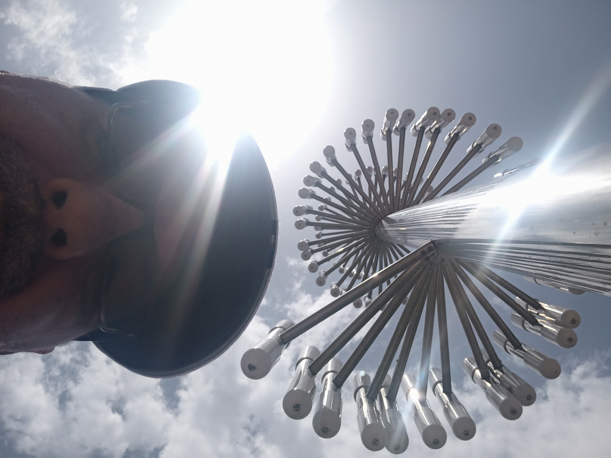 A low-angle, wide shot captures a person's face and a distinctive metallic sculpture against a bright, partly cloudy sky. The person, viewed from below, wears a dark cap and glasses, with a mustache visible. The sculpture is a tall, cylindrical structure with numerous metallic rods radiating outwards, each ending in a silver bulbous tip. The sun is bright, creating lens flare and obscuring details in some areas of the image. The photograph appears to be taken outdoors, possibly in a public space or park, with the sculpture being a prominent feature. The image does not provide explicit information about the location within the city of Dushanbe, Tajikistan.