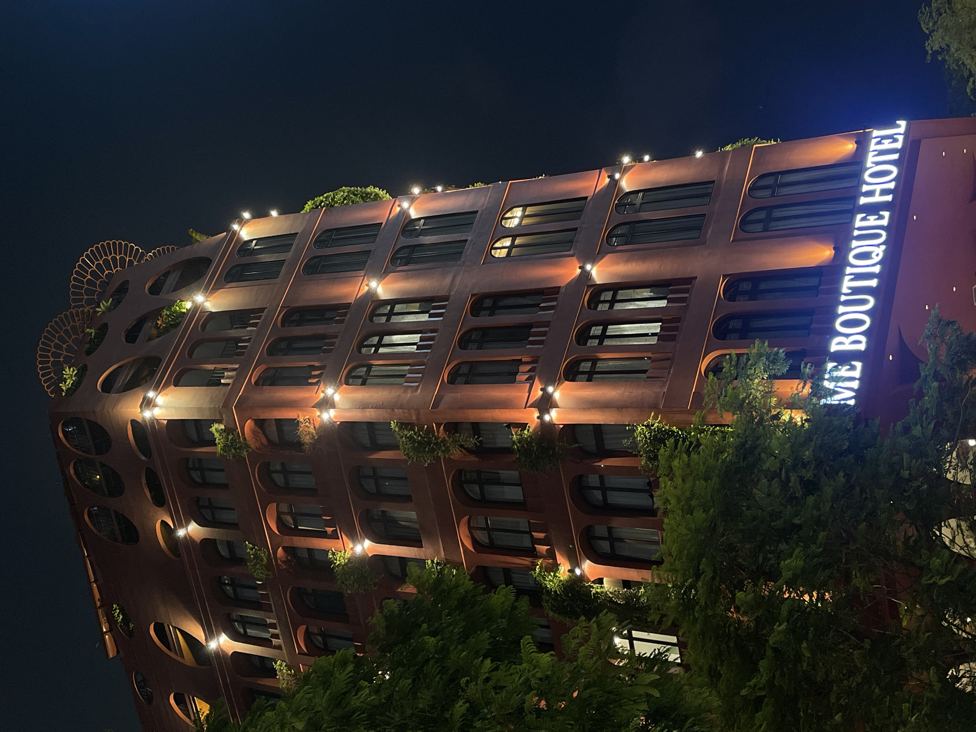 A night shot depicts a distinctive red-brown boutique hotel with multiple stories in Thủ Đức, Vietnam. The building features rounded window frames, rows of lights along its facade, and vegetation on some levels. A bright sign on the side reads "ME BOUTIQUE HOTEL." Trees obscure portions of the lower building levels. The sky is dark.
