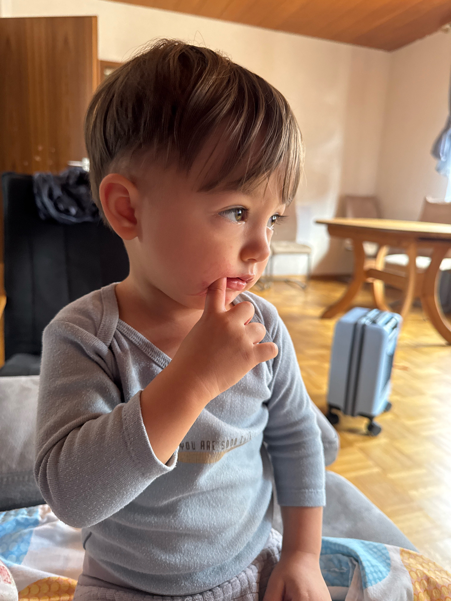 A toddler with brown hair is being photographed indoors, looking to the right with their finger on their mouth. The child is wearing a light grey long-sleeved shirt and grey pants. The background shows a room with wooden flooring, a wooden table, a blue suitcase, and a door. The location is identified as Überherrn, Germany.