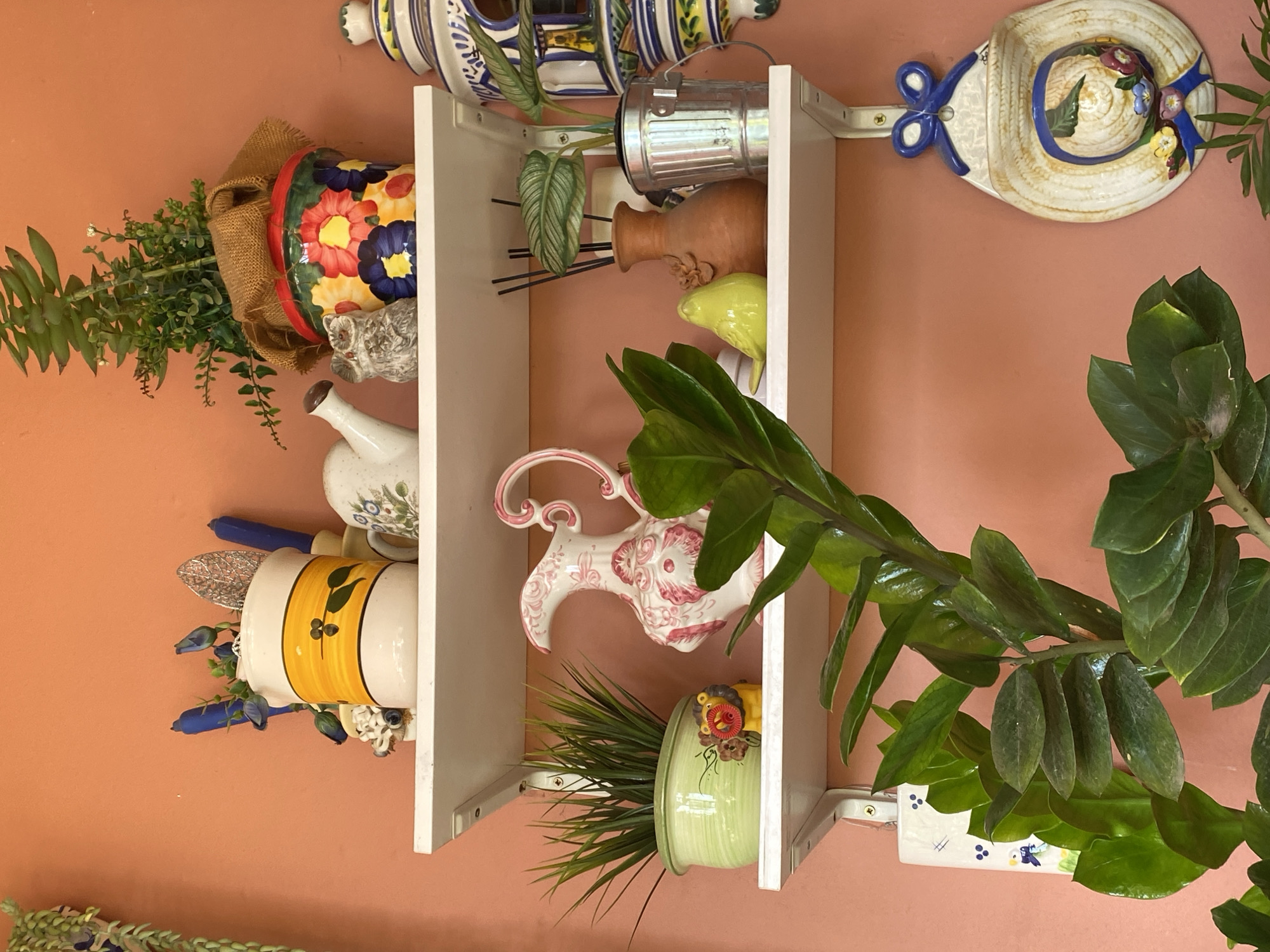 This is a medium shot, eye-level, indoor photograph of a wall with three white shelves. The wall is painted a solid, peachy-orange color. The shelves are decorated with various ceramic objects and plants. On the top shelf, there is a white ceramic pitcher with a yellow band and a black leaf design, a white ceramic watering can with a floral pattern, and a colorful ceramic planter with a plant. There are also a few blue candles and a small owl figurine. The middle shelf holds a green potted plant with grass-like leaves, a small yellow lion figurine, a pink and white patterned pitcher, a small yellow bird figurine, a terracotta vase with reeds, and a silver bucket with a plant. The bottom shelf is mostly obscured by a large plant with thick, glossy green leaves in the foreground. To the right of the shelves, a ceramic wall hanging shaped like a straw hat with a blue ribbon and flowers is attached to the wall. The scene is well-lit, with natural light coming from the left. The overall impression is one of a decorated indoor space, possibly a home or a shop, showcasing decorative ceramic items and greenery. The location in Resinera-voladilla, Spain is provided contextually.