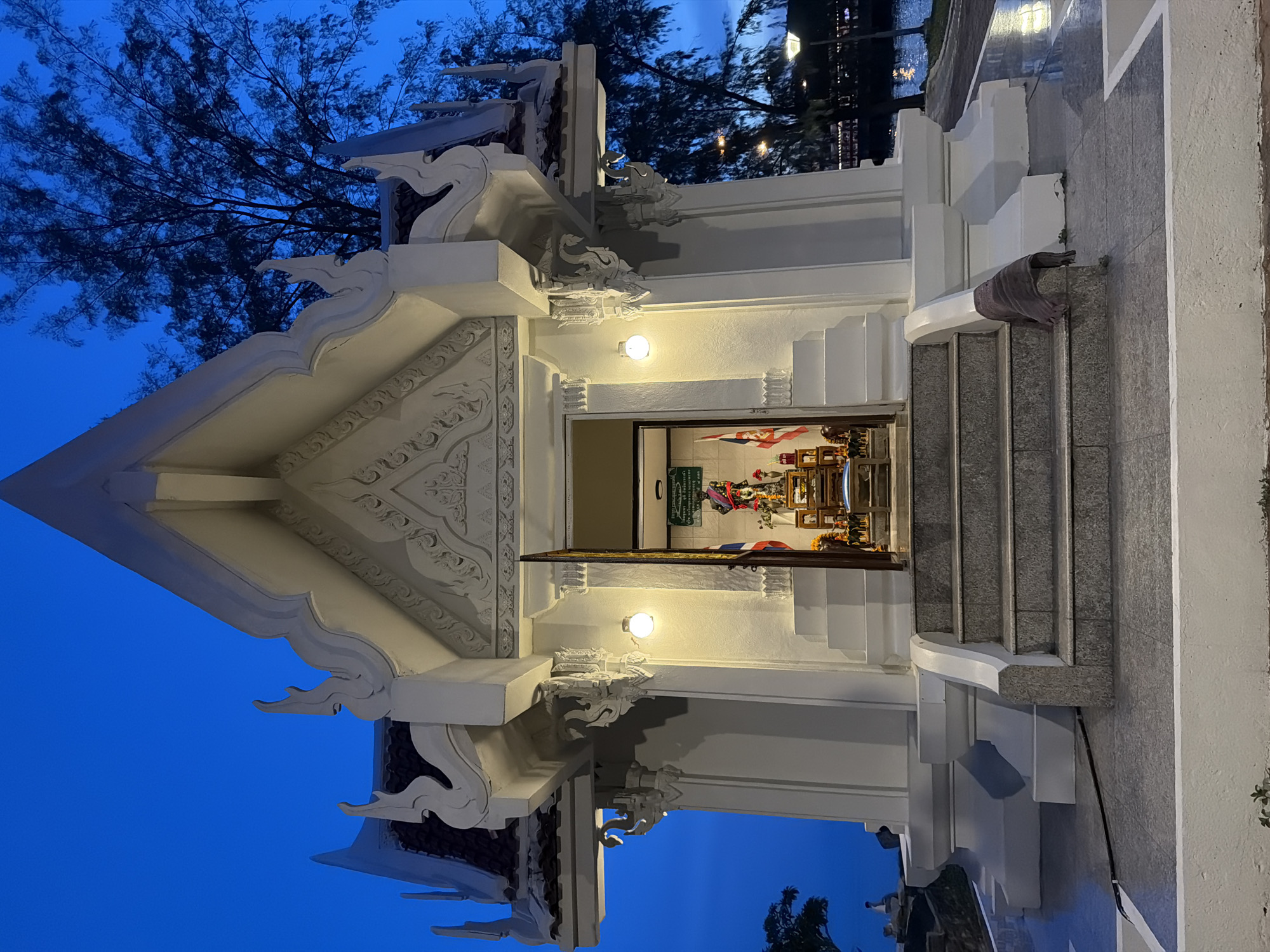 This is a medium shot, eye-level, outdoor, full-body photograph of a small white shrine with a gray stone staircase leading up to its entrance. The shrine features traditional Thai architectural elements, including ornate eaves and decorative carvings on its facade. The entrance is open, revealing a small altar with a statue of a deity adorned with red and gold fabric, surrounded by offerings and two small flags. Several lights illuminate the shrine from the exterior, and two lights are visible inside.
The shrine is situated on a paved area with a walkway to its right, which leads to a body of water, possibly a canal or a bay, reflecting the lights from the opposite bank. A large tree with sparse foliage stands to the right of the shrine, silhouetted against the deep blue twilight sky. In the distance, on the left side of the image, a person can be seen sitting near the water's edge, and a tree-covered hill is visible. The ground in front of the shrine is paved with gray stone tiles. A dark gray towel or cloth is draped over the railing of the staircase. The scene is lit by artificial lights and the ambient light of dusk, creating a serene and somewhat mystical atmosphere.