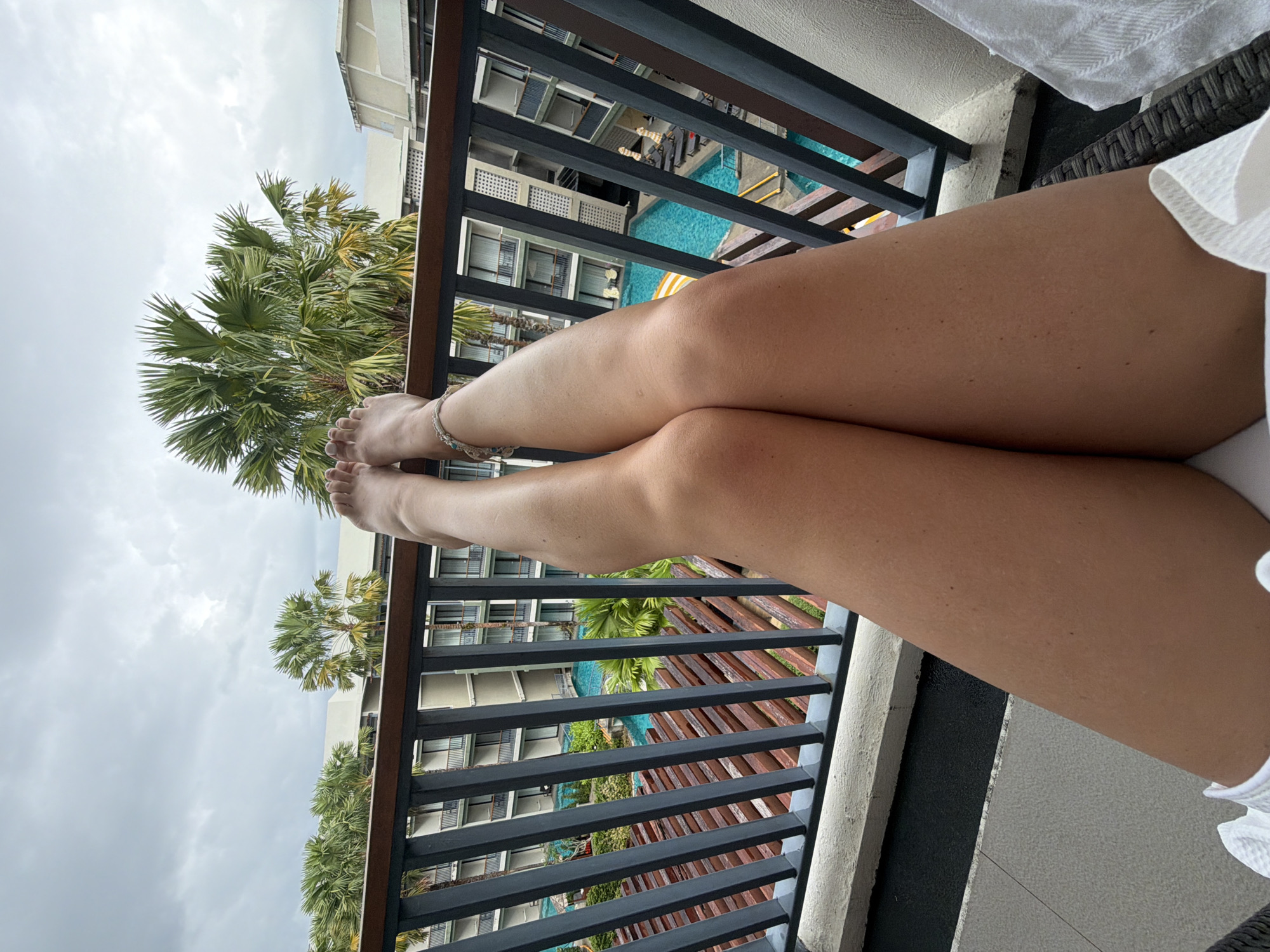 The image shows a first-person perspective of a person's legs resting on a balcony overlooking a resort in Bang Muang, Thailand. The legs are crossed at the knees and bare, with a beaded anklet visible on the left ankle. The balcony has a dark railing and a white concrete ledge. Beyond the railing, palm trees and a multi-story resort building with blue swimming pools are visible. The sky is overcast.