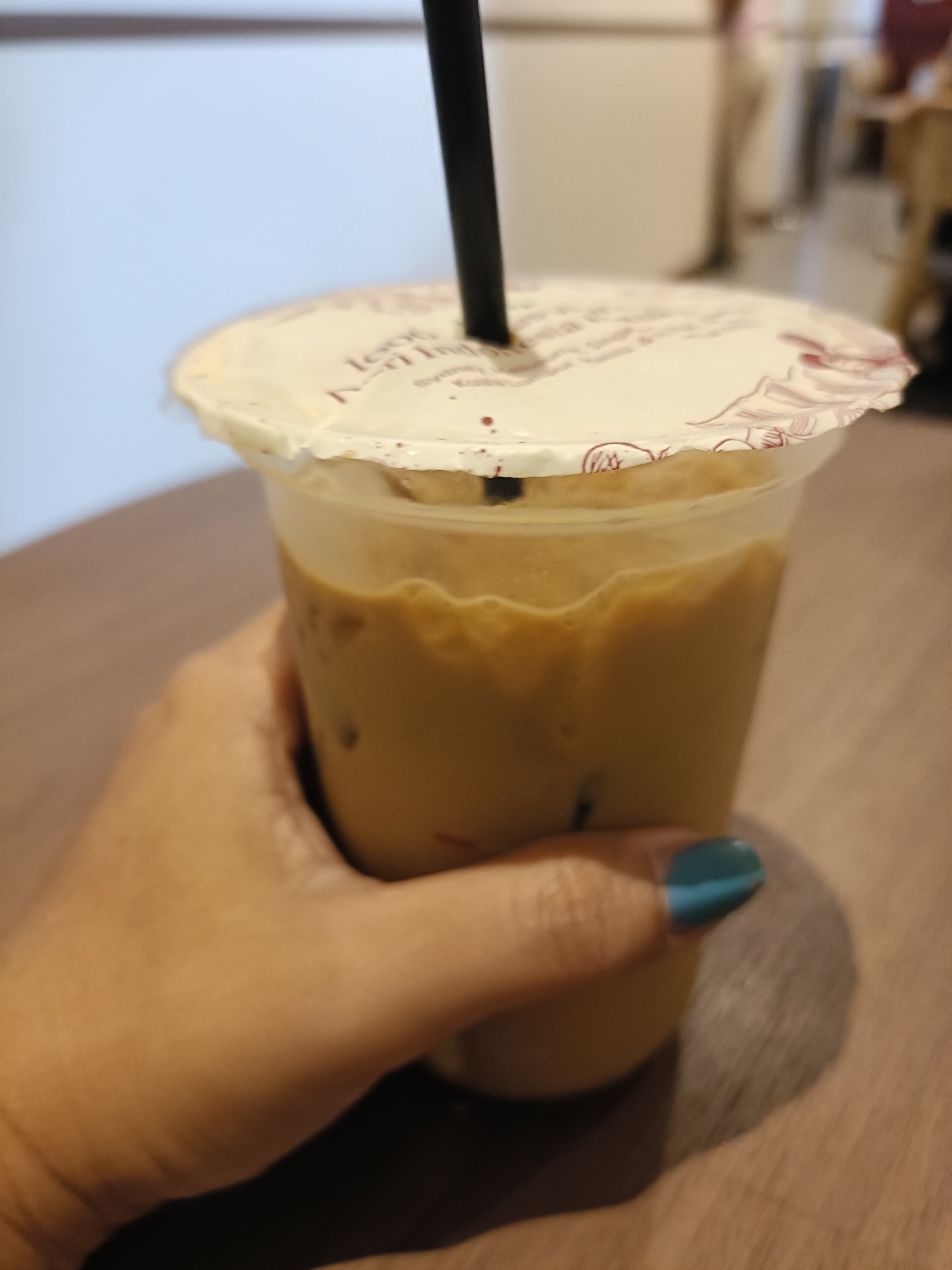 A close-up shot shows a hand holding a plastic cup of iced coffee. The cup has a white lid with some text and decorative elements printed on it. A black straw is inserted through the lid into the drink. The coffee is a light brown color with visible ice cubes. The background is blurred and appears to be an indoor setting, possibly a cafe or restaurant, with a wooden surface in the foreground. The image does not contain enough information to determine the location as Semarang, Indonesia.
