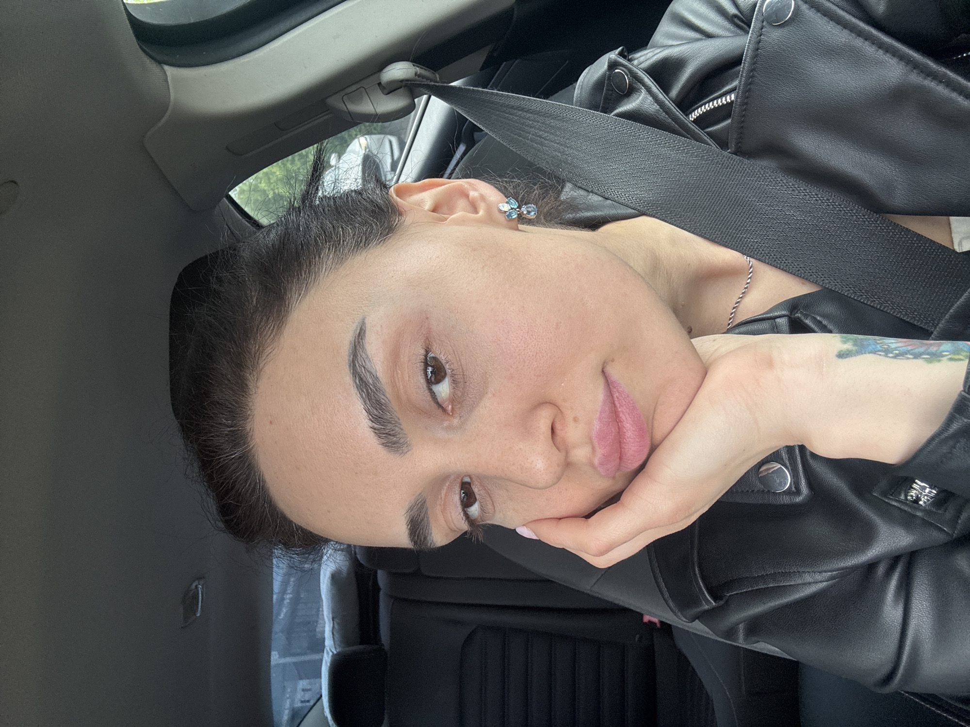 A woman is in a car, wearing a black leather jacket and a seatbelt. Her head is resting on her hand. She has dark hair pulled back, a silver necklace, and earrings. The car's interior is visible, including the headliner, seat, and window. The location is not identifiable from the image.