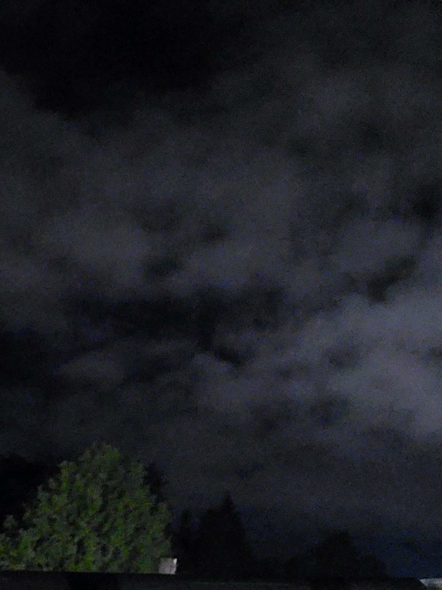 The image shows a nighttime sky filled with dark, textured clouds. In the lower portion of the image, the silhouettes of trees and the top of what appears to be a building are visible against the dark sky. The overall scene is dim and somewhat indistinct due to the low light conditions. The location is identified as Ozolnieki, Latvia.
