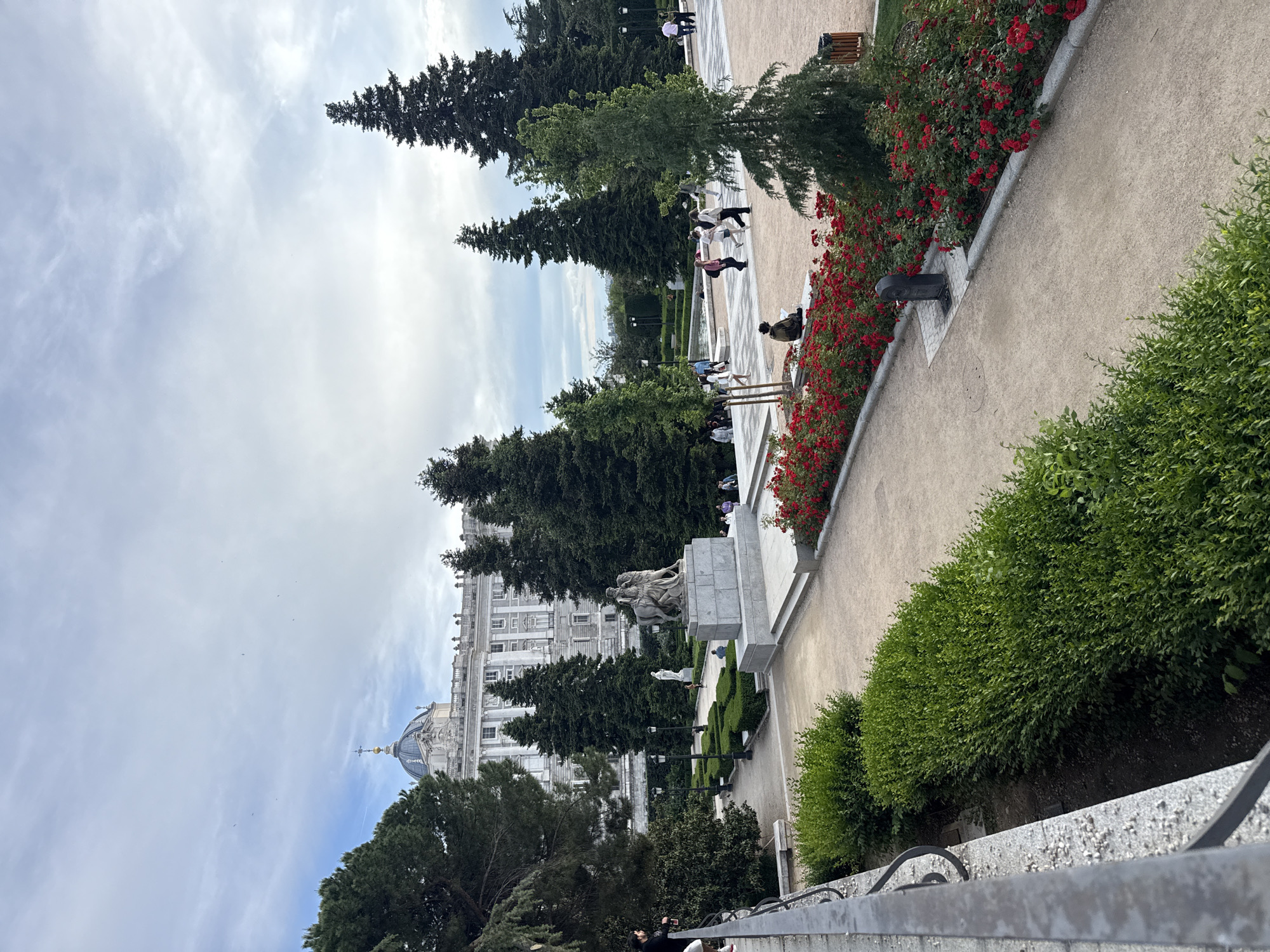 The image captures a scenic view in Madrid, Spain, possibly within the grounds of the Royal Palace or a nearby park. The Royal Palace is visible in the background. The scene includes pathways, flowerbeds with red flowers, trees, statues, and manicured hedges. People are seen strolling along the pathways. A sculpture of a man riding a horse is visible in the foreground. The sky is cloudy, suggesting fair weather.