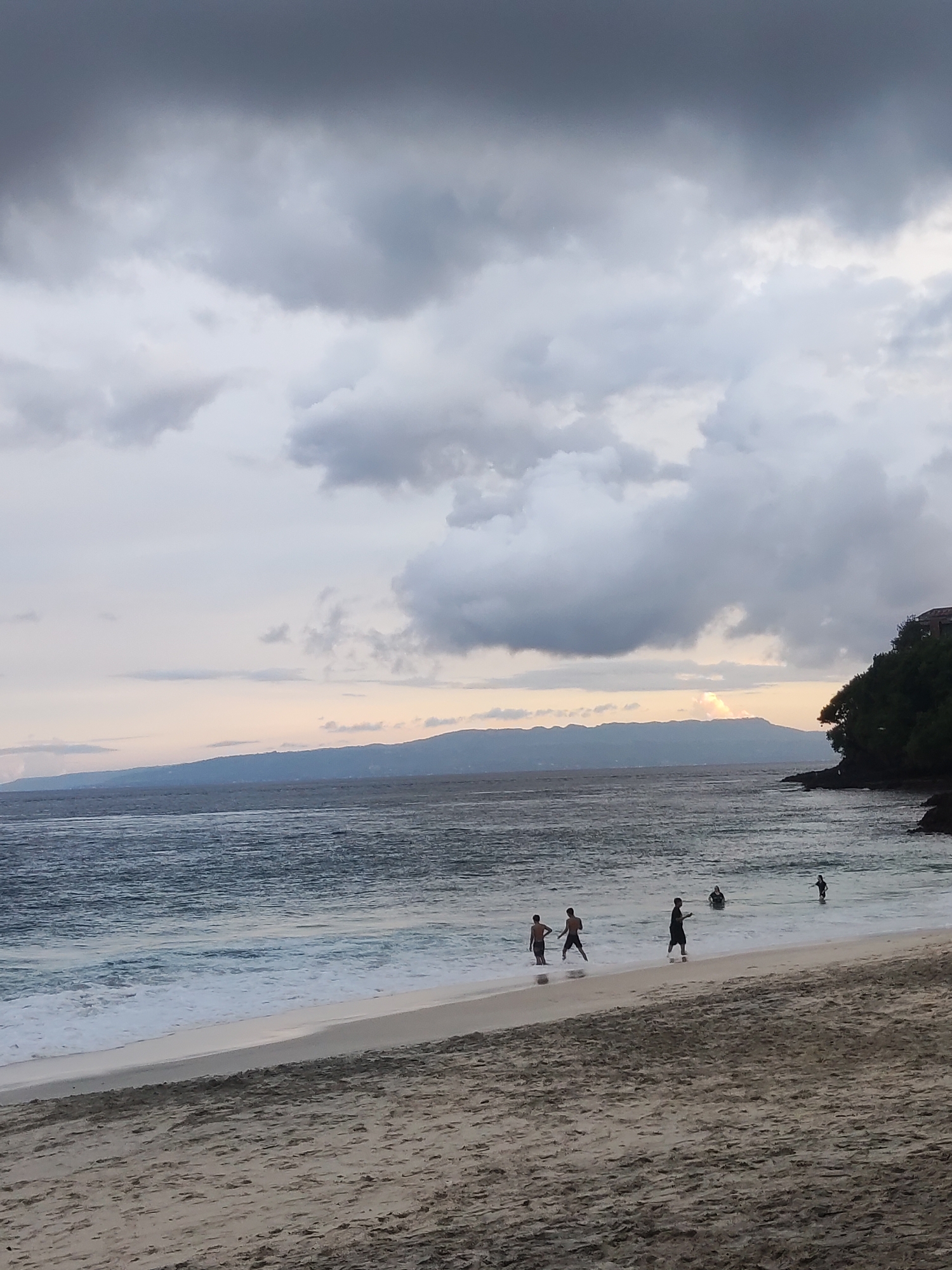 The image depicts Padangbai Beach in Indonesia. The sky is overcast with dark, heavy clouds dominating the upper portion of the frame. A sliver of sunlight pierces through the clouds near the horizon. The sea is moderately choppy with visible waves breaking along the shoreline. Five figures are present on the beach and in the water. The beach is sandy, with visible footprints. A vegetated landmass juts into the sea on the right side of the frame. Mountains are visible in the background across the water.