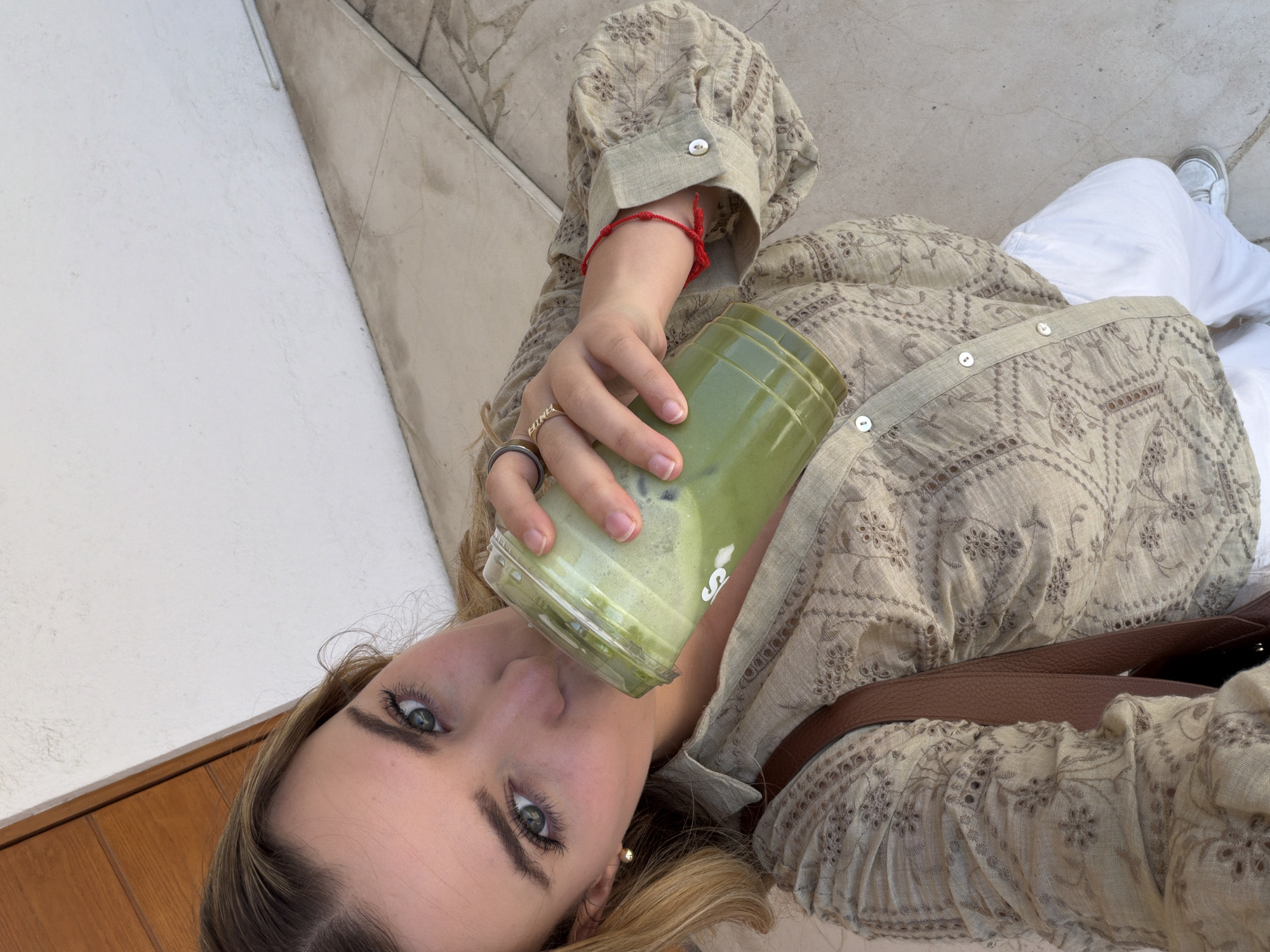 A woman is holding and drinking a green beverage from a clear plastic cup. She wears a tan patterned blouse with long sleeves and a brown shoulder bag. Her pants are white. She is also wearing a red bracelet on her left wrist and several rings on her left hand. The surrounding environment consists of a white wall and concrete steps, possibly located in an urban setting. The location in the city of San Bernardino Tlaxcalancingo, Mexico, is not discernible from the image.