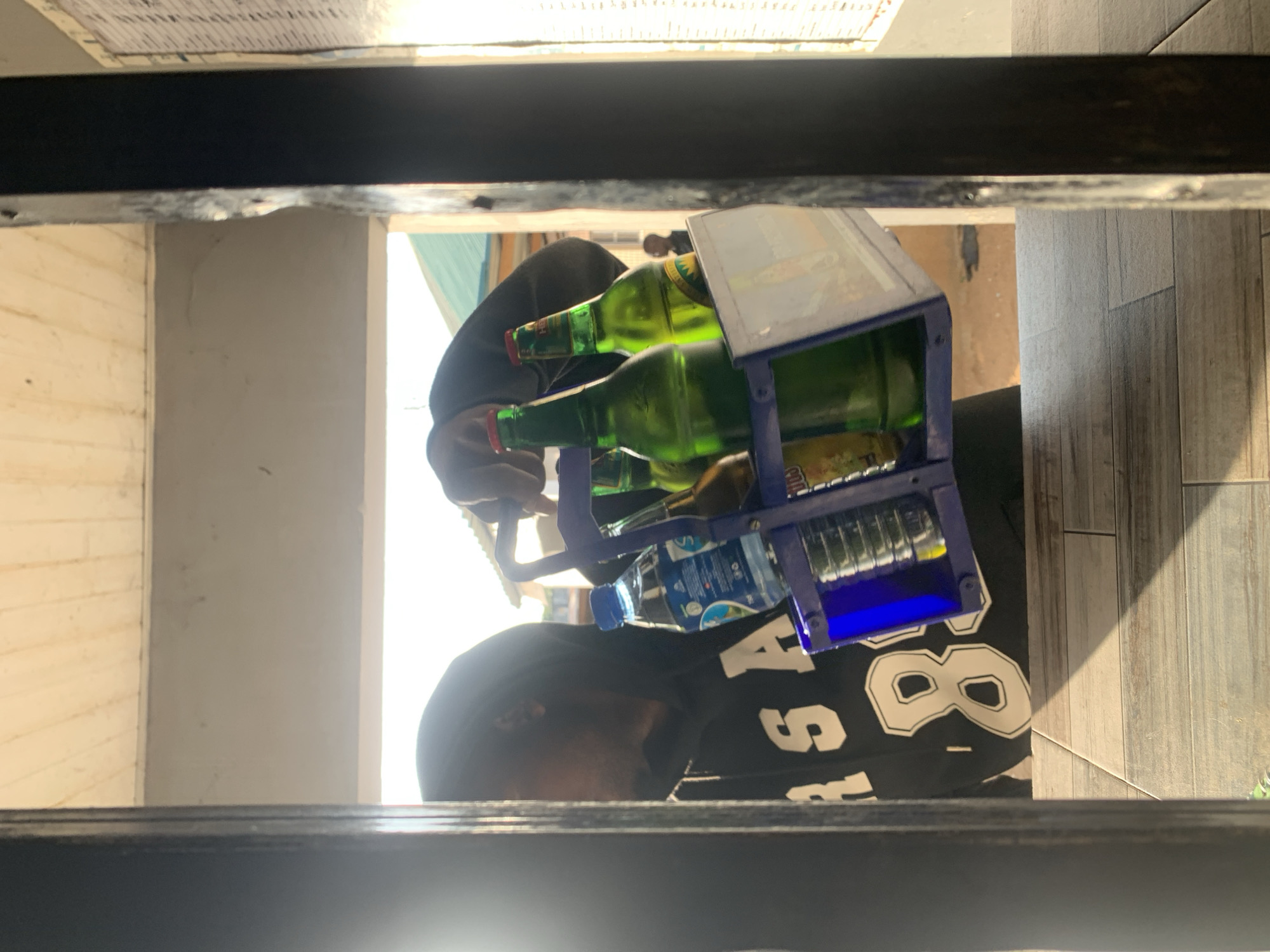 The image depicts a scene in New Stadium, Nigeria, viewed from an interior space looking outwards through an opening framed by dark vertical structures. In the immediate foreground, a tiled surface with a plank-like pattern is visible, marked by a sharp diagonal shadow extending from the right.
A partially visible individual, obscured by shadow and a dark hooded sweatshirt with white lettering "RS A" and the number "8", occupies the left side of the frame. This individual's right hand holds a blue plastic bottle carrier featuring a handle. The carrier contains one clear plastic water bottle with a blue cap and a partially legible "AQUA S" label, along with at least two green glass bottles with red caps, and one partially visible brown bottle. A small, illegible advertisement panel is integrated into the front of the carrier.
The background is brightly illuminated by daylight. Above the individual and carrier, a light-colored wall or ceiling segment is visible, topped by a slatted wooden ceiling structure. To the right of the bottle carrier, a faint silhouette of a second person is discernible against the bright light. Further right, a vertical panel with dense black text or markings, resembling a ledger or notice board, is strongly illuminated. A greenish-blue corrugated roof structure is present in the mid-distance on the right. The ground beneath the bottle carrier appears to be reddish-brown earth.