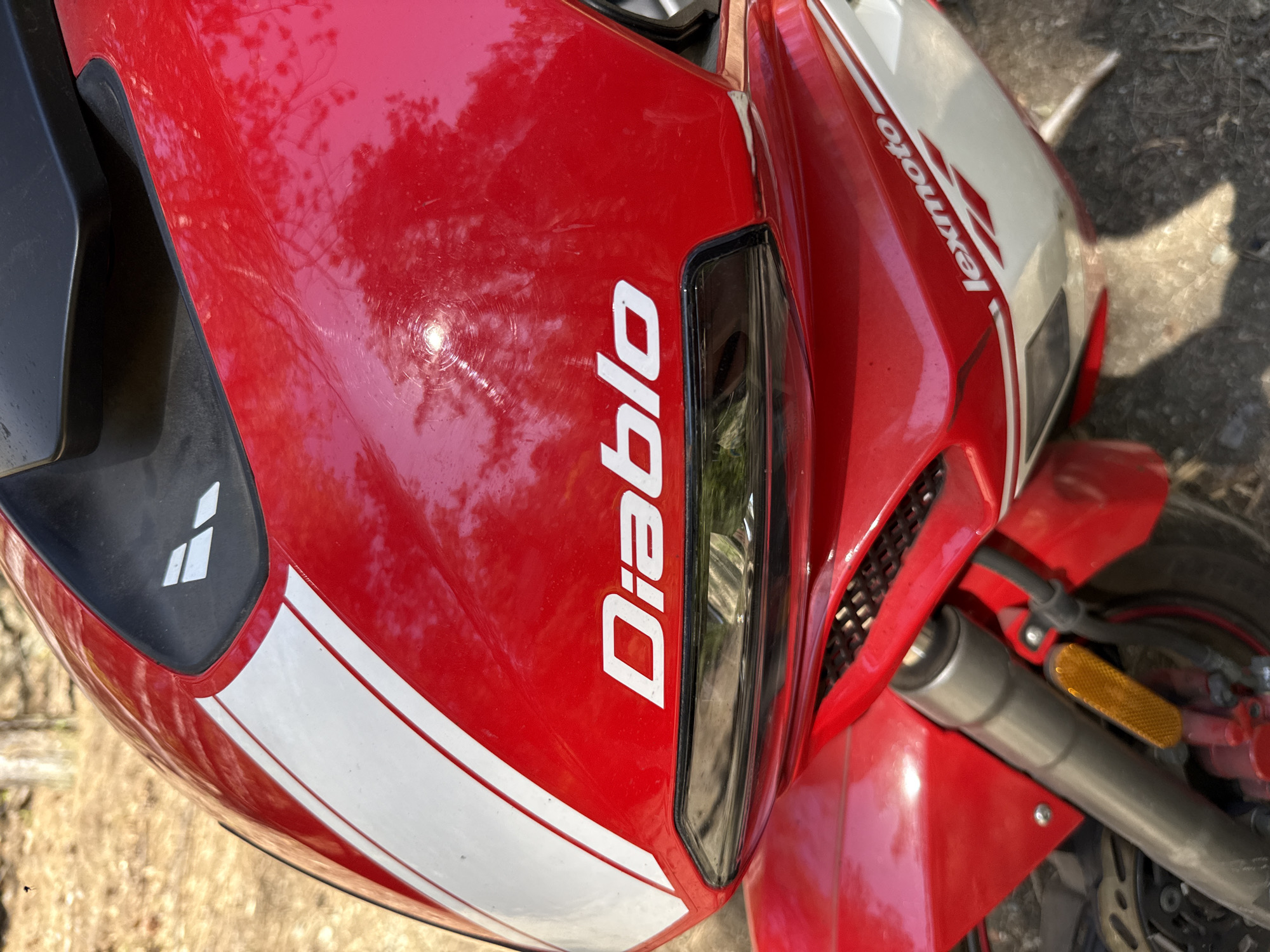 The image shows a portion of a red Lexmoto Diablo motorcycle. The word "Diablo" is printed vertically in white on the red fairing. The front headlight is visible, along with white stripes on the fairing. Part of the front wheel and suspension are in the lower right of the frame. The location is not evident from the image.