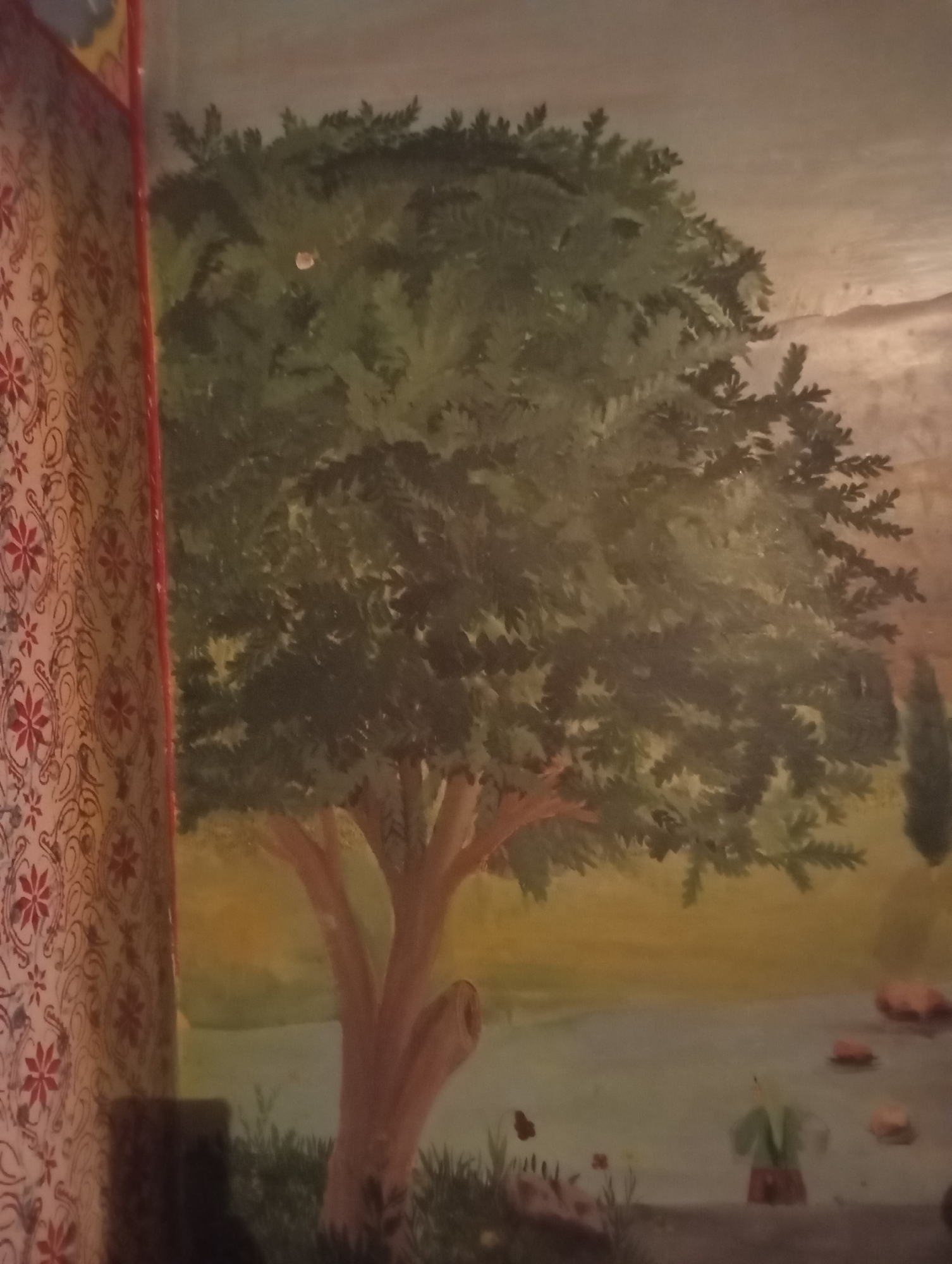 The image depicts a mural painting, likely on an interior wall, in Samarqand, Uzbekistan. The mural features a large, leafy tree as the central subject, with its trunk and branches rendered in shades of brown and green. The background suggests a landscape with a body of water and distant hills or mountains, painted in muted earthy tones. To the left, a section of patterned fabric, possibly a curtain or tapestry, with red floral motifs on a light background, partially obscures the mural. In the lower right portion of the mural, a small, stylized figure wearing green and red is visible near the water, accompanied by several rocks or pebbles. A butterfly is also depicted in flight near the foreground grass. The overall style appears to be illustrative, possibly reflecting traditional Uzbek artistic themes.