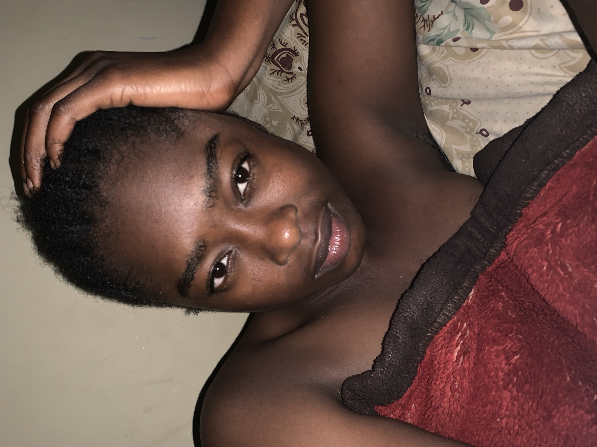 A close-up of a dark-skinned person with short hair, lying down on a surface covered with bedding, and partially covered with a red and black blanket. The person's hand is on their head. The background includes a wall. The location of the image is unknown and there is no indication it is in Rafin Par, Nigeria.