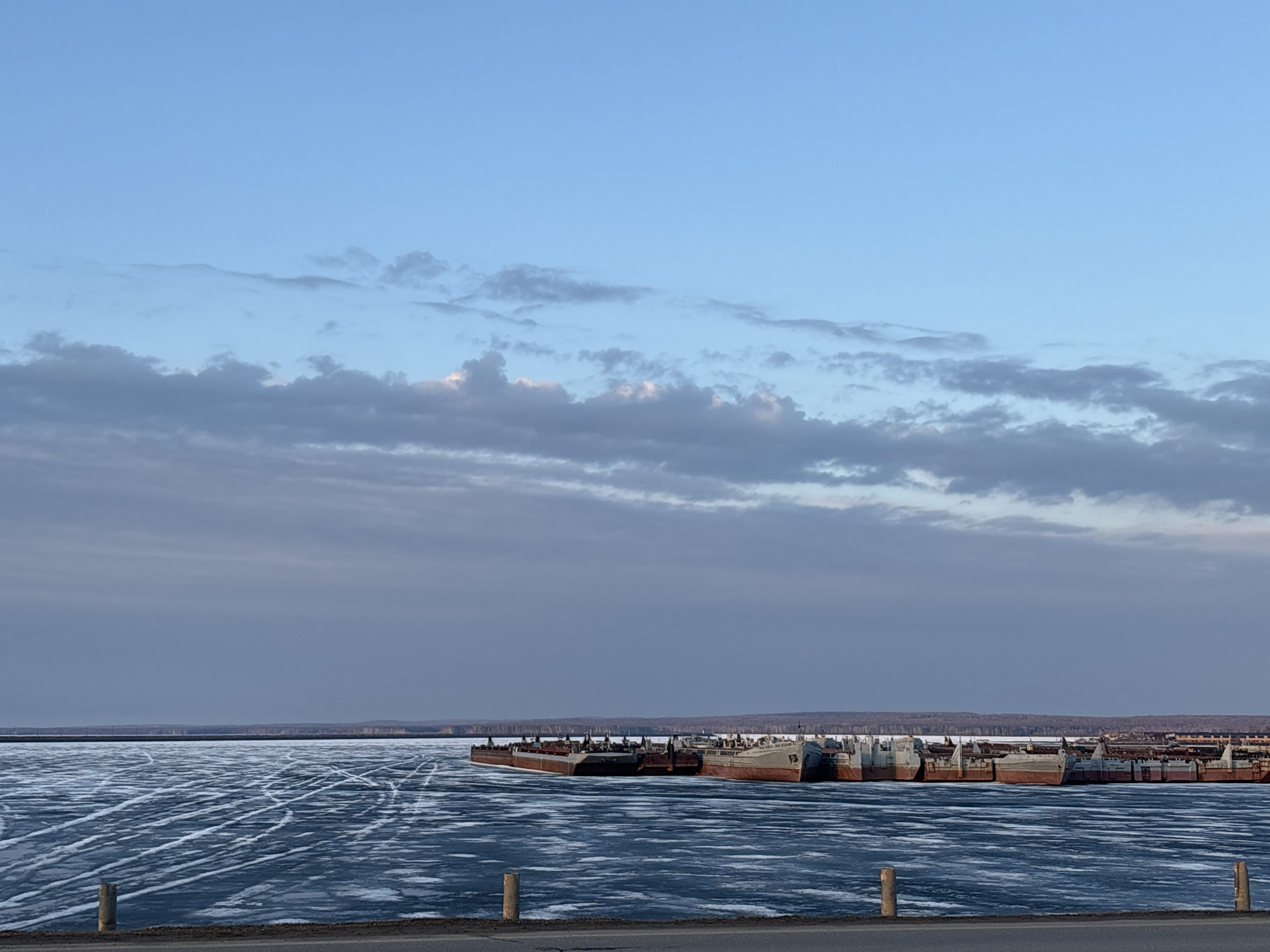 The image depicts a frozen body of water, potentially a river or reservoir, with numerous barges docked or grounded along the edge. The surface of the ice is marked with tracks, possibly from vehicles or foot traffic. In the background, a distant treeline is visible. The sky is overcast with a mix of light blue and grey clouds. The location is Chaikovsky, Russia.