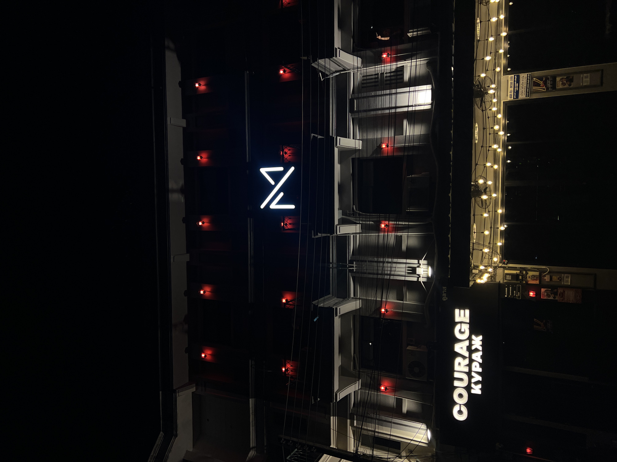 Captured at night in Unknown, Unknown, the image presents a building facade lit with a mixture of light sources. A white neon sign displaying a stylized "Z" takes center stage, flanked by rows of smaller red lights accentuating the structure's edges. A vertical sign spelling "COURAGE" in bold, white letters, accompanied by "КУРАЖ" in Cyrillic, appears on the lower right side of the image, indicating a business establishment. A string of warm-toned lights adorns the top right, adding to the eclectic illumination. The architectural details include ledges, windows, and external fixtures, emphasizing the building's multi-level construction.