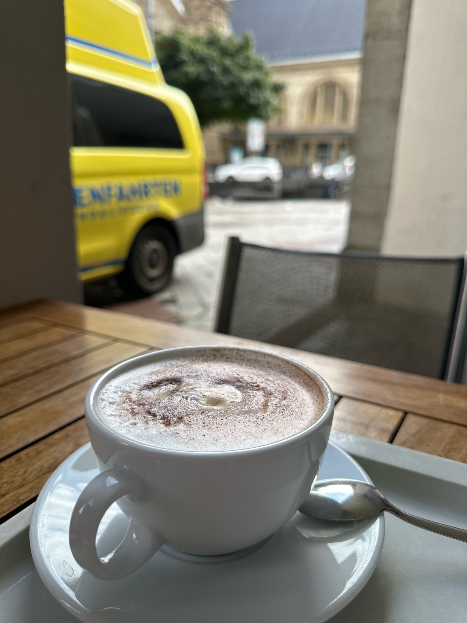 The image shows a close-up view of a white cup filled with a foamy beverage, likely hot chocolate or cappuccino, dusted with cocoa powder. The cup sits on a saucer, which is placed on a light gray tray. A silver spoon rests on the saucer, to the right of the cup. The setting appears to be an outdoor cafe, with a wooden table visible beneath the cup. In the background, out of focus, is a yellow van with blue stripes and lettering. Further in the distance, blurred buildings and trees suggest an urban environment. Based on the van's markings, which include "NOTFALLKRAFT", the scene is located in Krefeld, Germany, indicating it is likely near an emergency service vehicle.