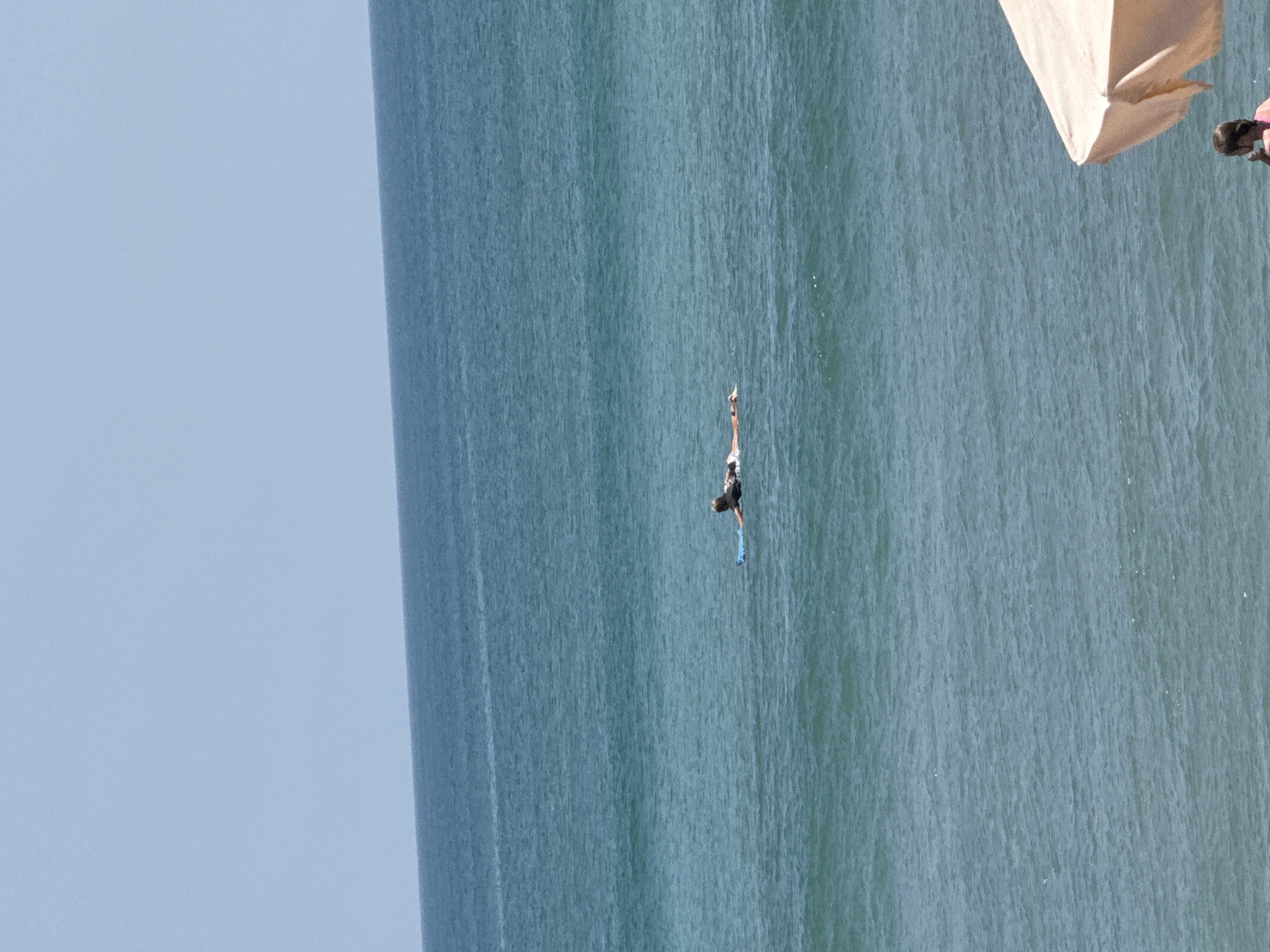 A person is seen lying on a blue surfboard in the ocean, with the water appearing calm and blue under a clear sky. In the foreground, a portion of a peach-colored beach umbrella is visible, with a person wearing a pink shirt seated underneath it. The location is Kuta, Indonesia.