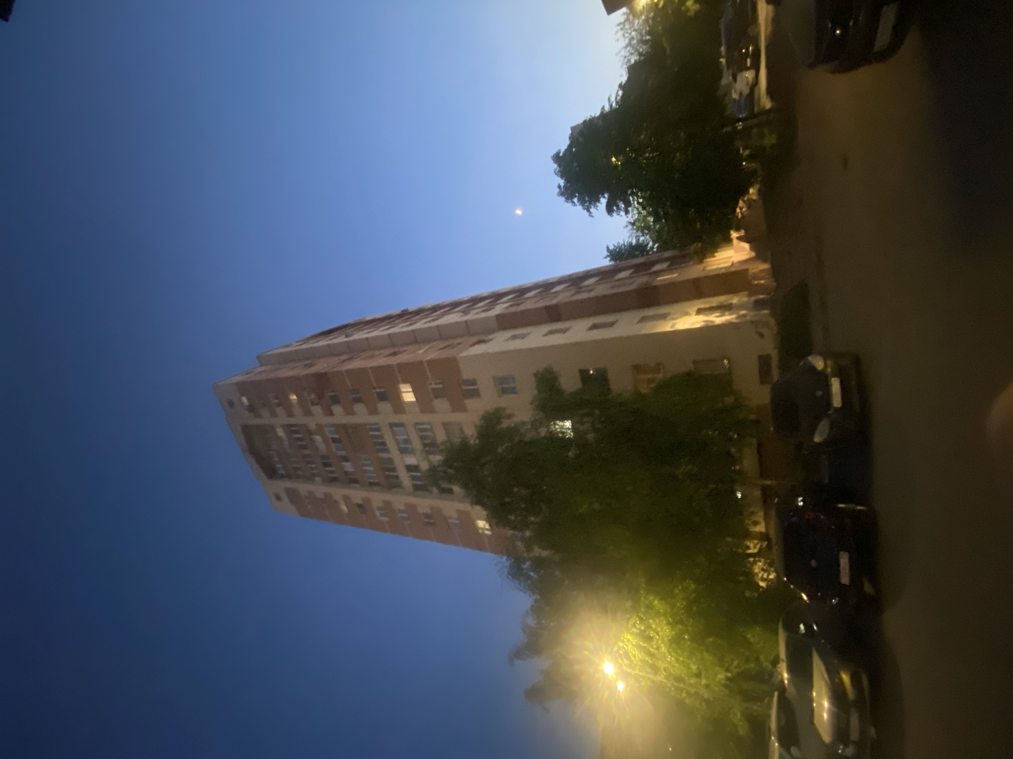 The image shows a tall, multi-story building in Kazan, Russia, likely an apartment complex. The building is primarily beige with reddish-brown accents. It is lit by warm interior lights. The sky transitions from a dark blue at the top left to a lighter blue near the building. Trees are present on the right side of the frame, partially obscuring the building. Streetlights are visible amongst the trees. A row of parked cars lines the right edge of the image, next to a dark, paved area that could be a street or parking lot. There is a faint crescent moon in the sky. There are no people visible in the image.