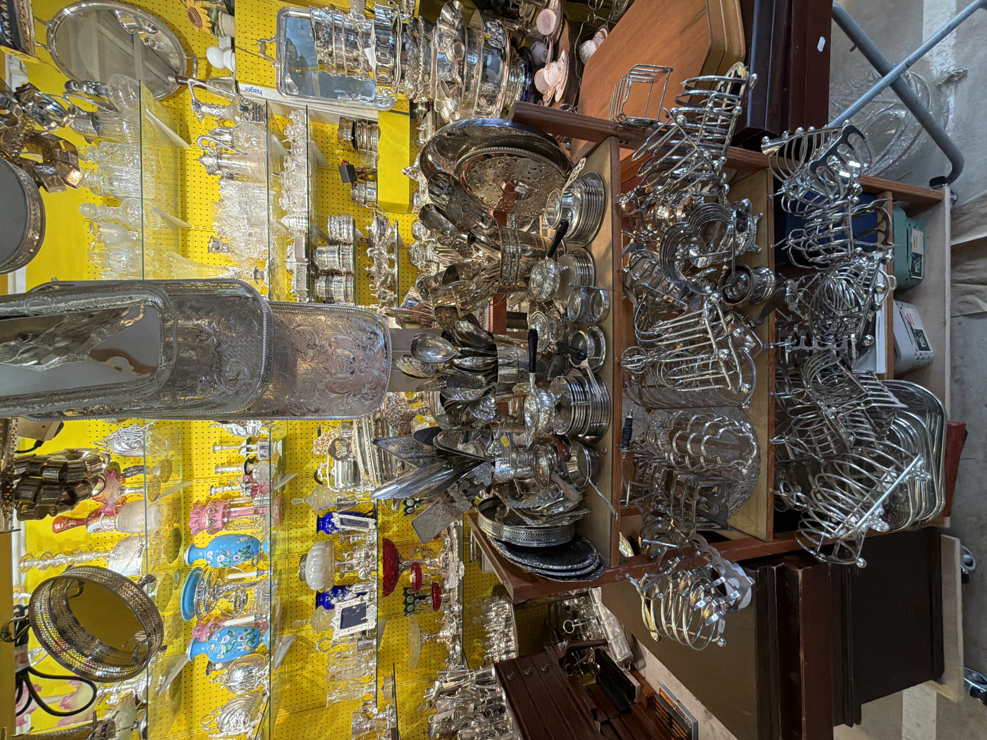 The image presents a cluttered display, possibly inside an antique or secondhand shop. Dominated by reflective silverware and glassware, the items are crammed onto shelves and within wooden cabinets. The background consists of a bright yellow pegboard, on which numerous glass objects and silver-plated items are hung. The shop's overall ambiance appears busy and filled with a diverse range of items, suggestive of a place offering a wide array of vintage or collectible pieces. I cannot provide the specific location in London based on the visual evidence alone.