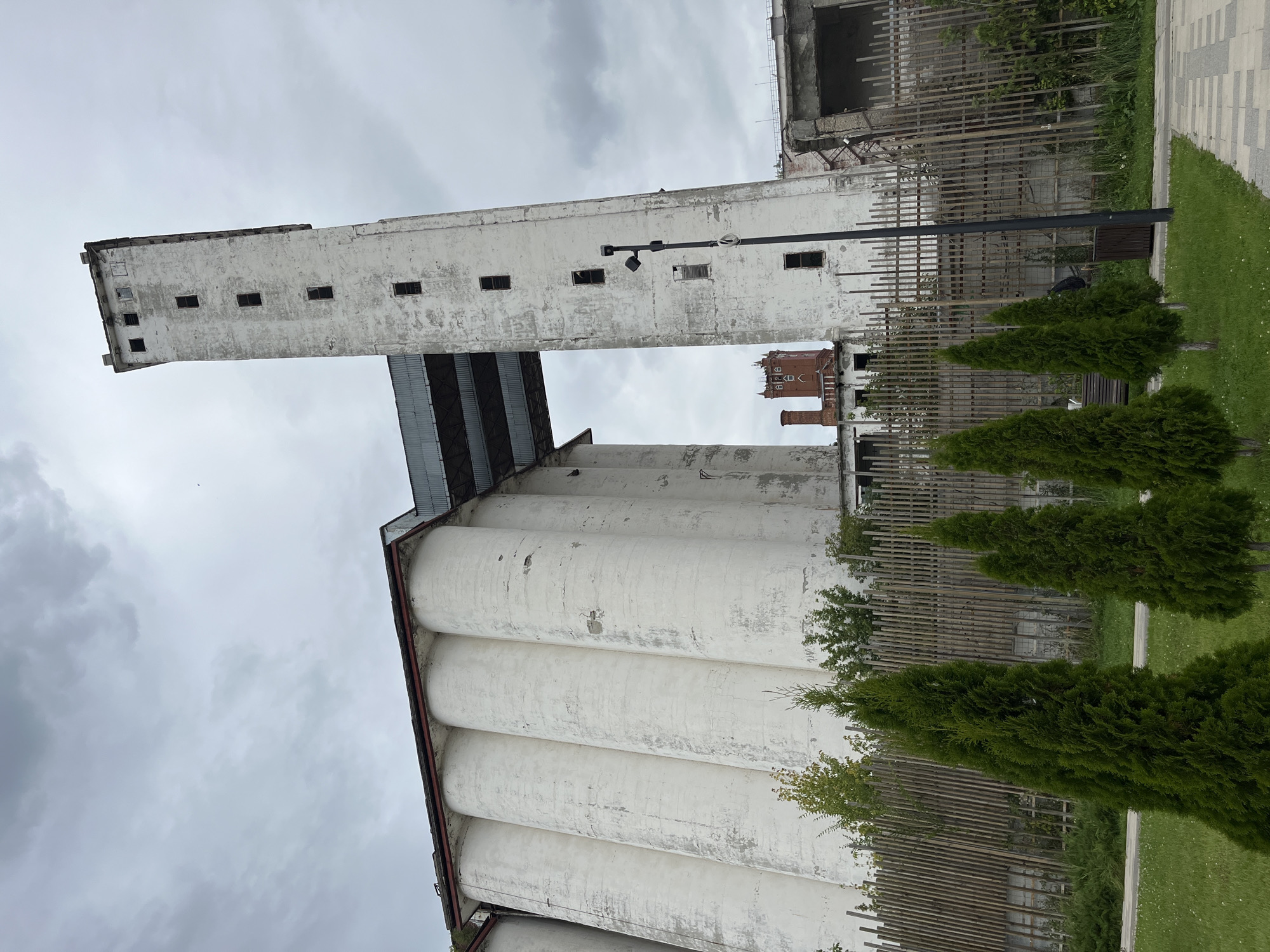 The image shows a view of the former Saratov grain elevator, an industrial complex located in Saratov, Russia. The structure is characterized by several large, white cylindrical silos connected by a covered walkway to a tall, white tower. The tower has numerous small, dark windows arranged vertically. The buildings appear weathered and show signs of age and disrepair, with peeling paint and some visible damage. In the foreground, a neatly maintained grassy area with manicured evergreen trees and a wooden fence separates the viewer from the industrial site. A paved walkway is visible on the right side of the image. The sky is overcast, with a cloudy, gray atmosphere. There are no people visible in the image.