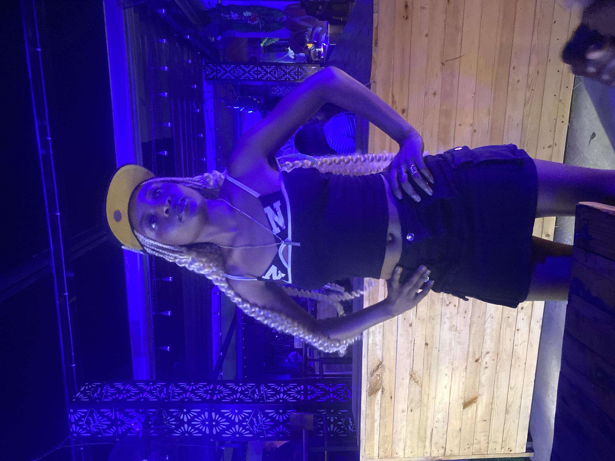 A young Black woman with blonde braids and a yellow baseball cap stands with her hands on her hips in front of a wooden wall. She is wearing a black crop top with white trim and a black skirt. The background is dimly lit with blue and purple lights, suggesting an indoor venue such as a nightclub or bar. There are decorative pillars with white patterns and lights in the background. A table is partially visible in the foreground, also illuminated by the blue and purple lights.