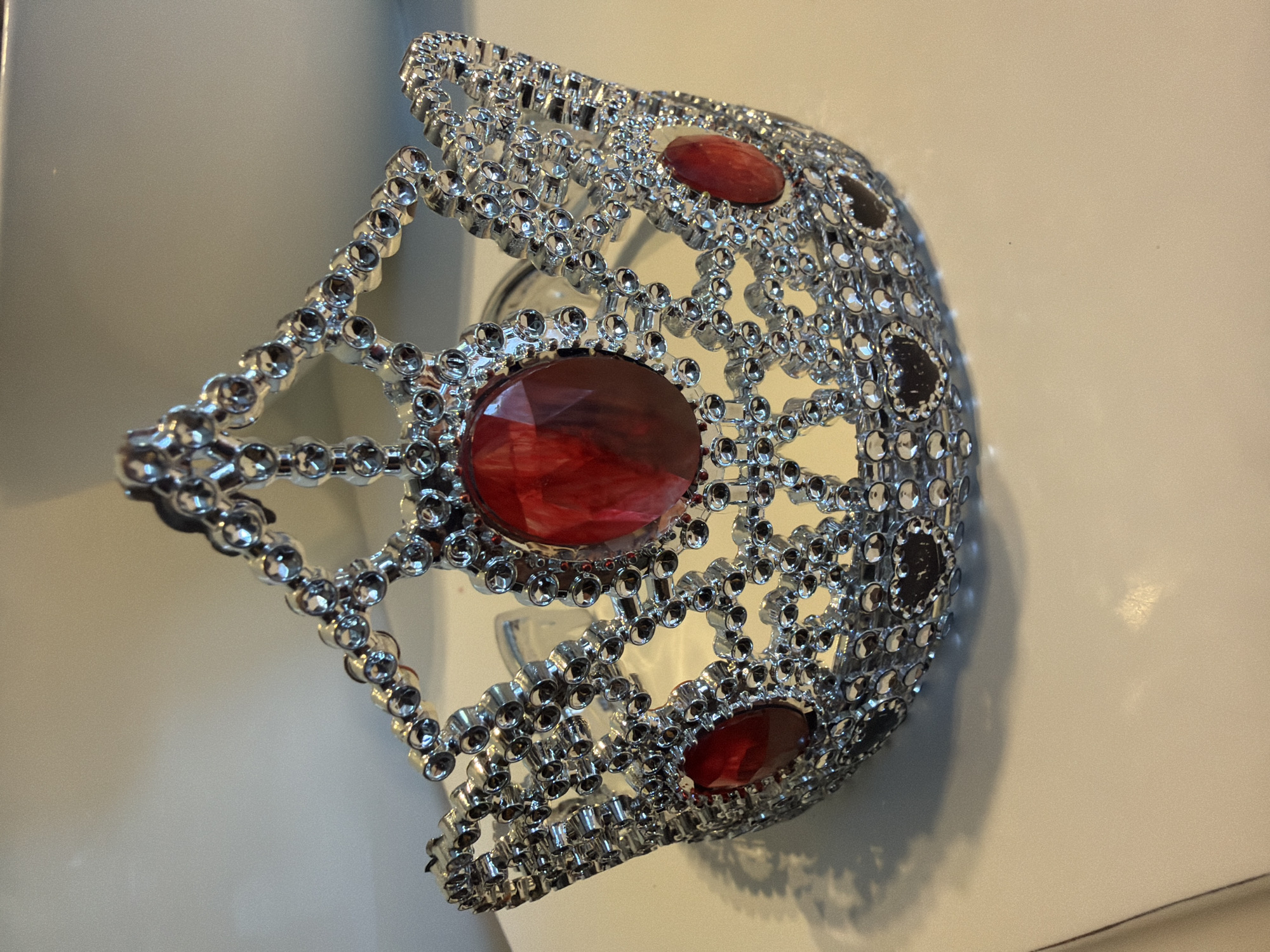 A decorative crown, silver-colored and adorned with numerous small, reflective beads and three prominent red oval jewels, is positioned on a light-colored, flat surface. The crown features an openwork, filigree-like design composed of linked circular elements. A large central red jewel is visible, flanked by two smaller red jewels positioned laterally. Dark, heart-shaped embellishments are present near the crown's base. The scene is depicted in the city of Baloži, Latvia, with a plain, light-toned background. No individuals or actions are present.