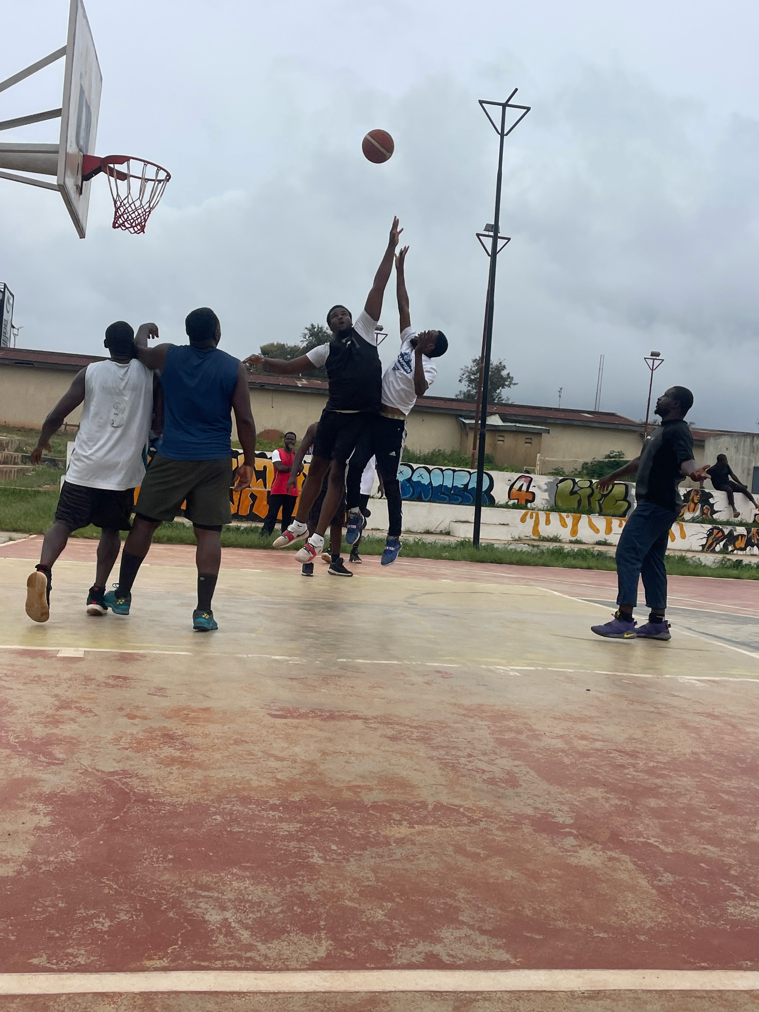 The image depicts a basketball game being played on an outdoor court in Jos, Nigeria, under an overcast sky. The court surface is a worn, reddish-brown concrete with white boundary lines. In the foreground, two young men are engaged in a jump ball, reaching for a basketball mid-air. One player, wearing a white sleeveless shirt and black shorts, is in the air with his arms extended upwards. The other player, in a black sleeveless shirt and dark shorts, is also airborne, his arms reaching for the ball.
Behind them, on the left side of the court, another young man in a white sleeveless shirt and black shorts stands with his back to the camera, observing the play. To his right, a young man in a navy blue sleeveless shirt and dark shorts is also watching the game. Further in the background, several other individuals are visible, some standing and others appearing to be involved in the game or spectating. A red and white basketball hoop with a net is positioned on the left side of the frame. Behind the court, a low concrete wall is covered in graffiti. In the distance, buildings and vegetation are visible, suggesting an urban or semi-urban environment. A tall, dark pole with a triangular fixture stands on the court near the center. The overall scene captures a moment of athletic activity on a community basketball court.