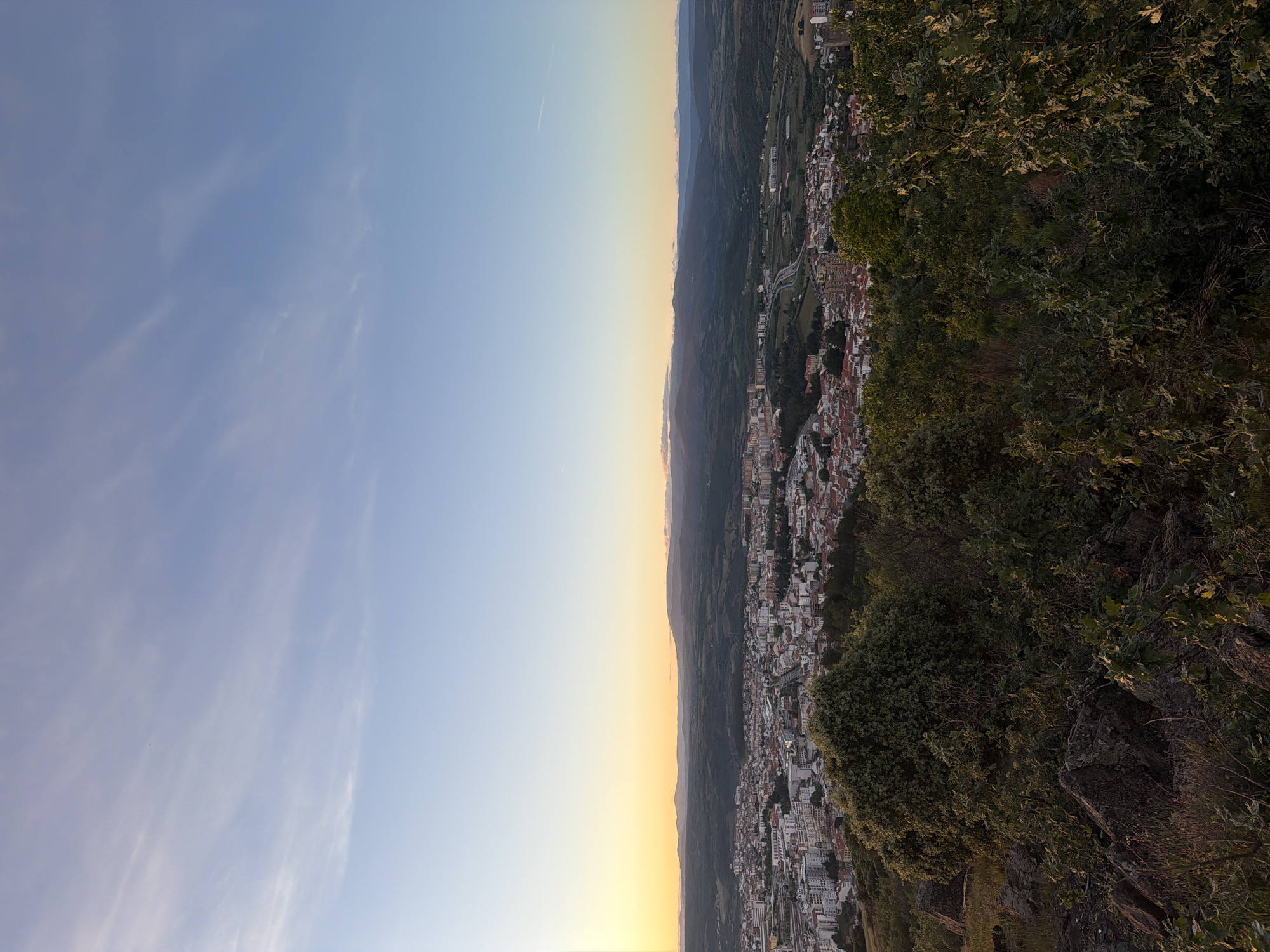 The image is a landscape photograph taken from a high vantage point, likely in the city of Bragança, Portugal. The photograph shows a view of a city nestled in a valley with rolling hills in the distance. The sky transitions from light blue to a yellow-orange hue, suggesting it is either sunrise or sunset. The right side of the image includes foliage and rocks, indicating the location is on a hill or mountain overlooking the city. The urban area includes buildings, roads, and possibly a river or stream. The overall impression is one of a scenic view of a city surrounded by natural beauty. There are no people visible in the image.