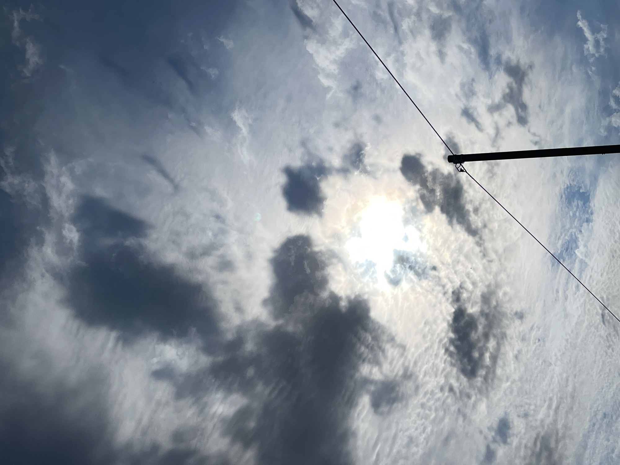 A vertical shot depicts a dusky sky filled with scattered clouds. In the lower right corner, a dark vertical pole, likely a utility pole, stands against the sky. A thin, dark wire stretches diagonally across the frame from the pole, extending towards the upper left. The sun is partially obscured by clouds, casting a bright glow in the central area of the sky. The overall impression is one of an overcast day with intermittent sunshine. The location of this scene is in the city of Donskoye, Russia.