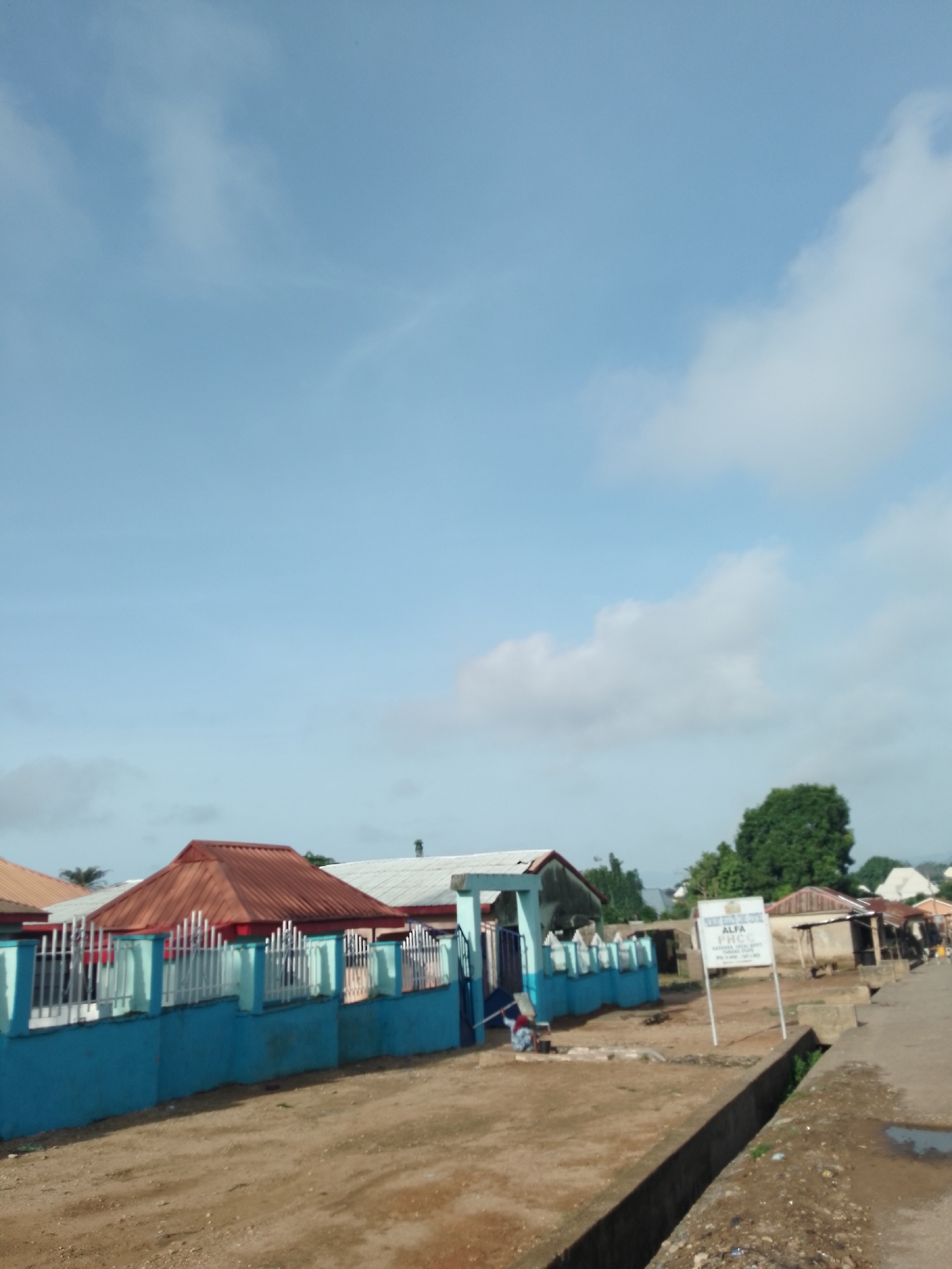 The image depicts a street scene in Serti, Nigeria. In the foreground, a dirt road runs alongside a concrete drainage channel. On the left side of the road, a blue wall with a white metal fence runs along a property line. A building with a reddish-brown, gabled roof is visible behind the fence. Further down the road, a white sign with black text stands on a metal pole. The sign appears to be for "ALFA PHC". Beyond the sign, several small structures and a large tree are visible. The sky above is a light blue with scattered white clouds. The overall impression is of a dusty, somewhat sparsely populated area.