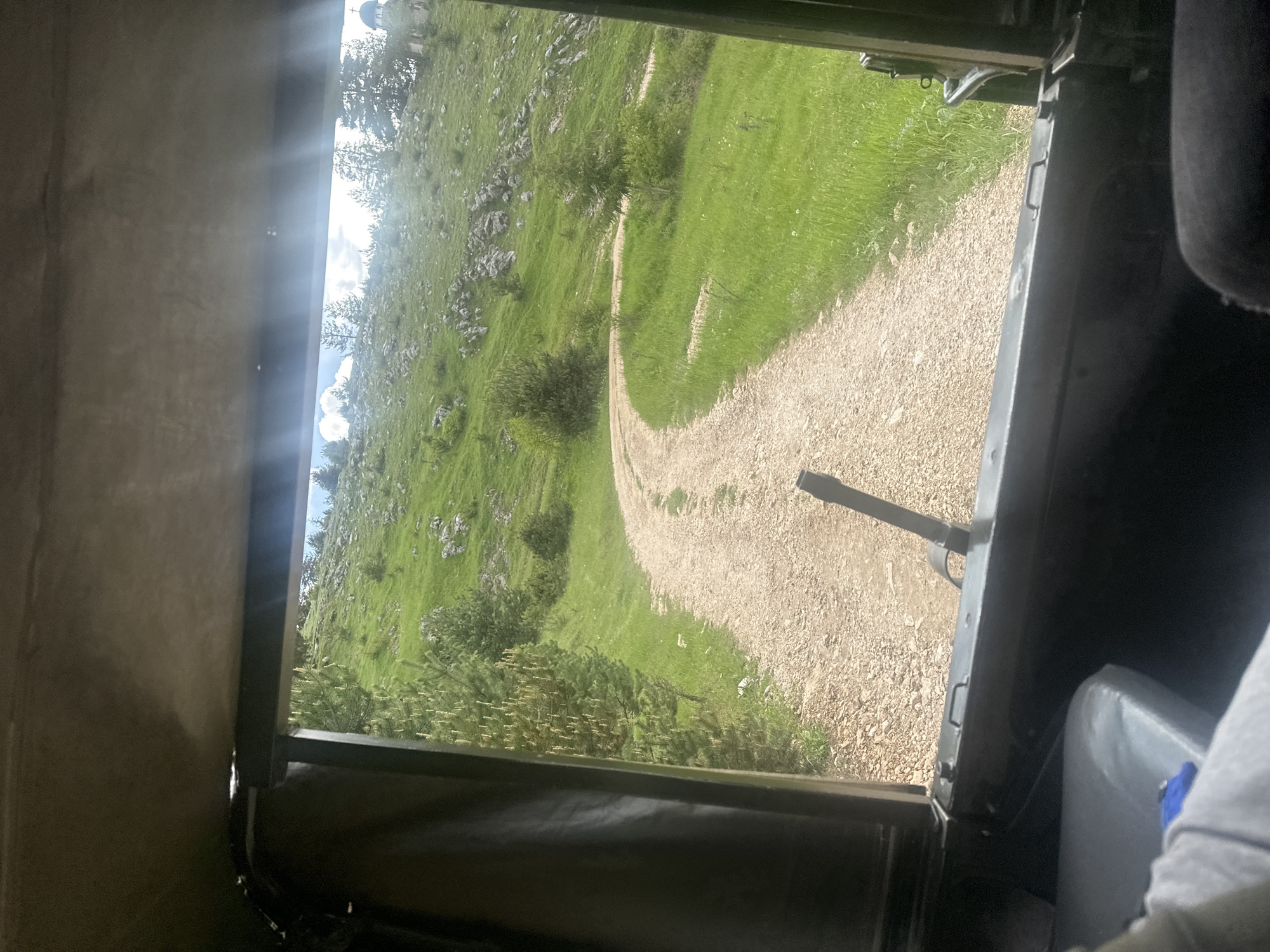 The view is from inside a vehicle, looking out at a gravel road winding through a grassy, rocky hillside. The hillside is dotted with trees and vegetation. The vehicle's interior is visible, including part of a seat. Bright sunlight illuminates the scene. The location cannot be determined from the image. Iagodina, Bulgaria, cannot be confirmed.