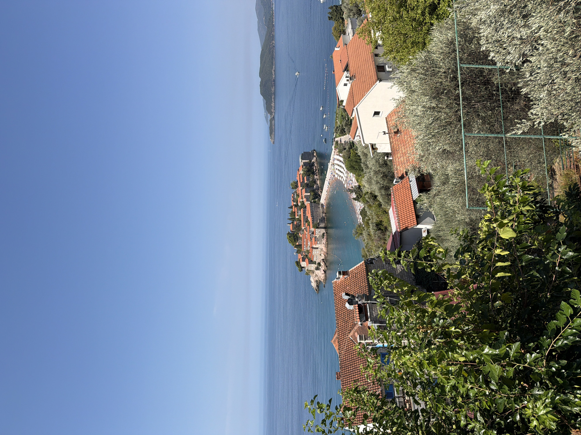 The image captures a scenic view of Sveti Stefan, Montenegro, under a clear blue sky. In the foreground, dense green foliage partially obscures a cluster of buildings with terracotta roofs. Beyond these structures, the azure waters of the Adriatic Sea stretch towards the horizon. A picturesque island, Sveti Stefan, is visible in the middle ground, characterized by its red-tiled roofs and stone buildings, connected to the mainland by a narrow causeway. A beach with numerous umbrellas is situated on the causeway and adjacent to the island. Several boats are dotted across the sea. In the background, lush green hills and a distant island rise from the water, further enhancing the tranquil coastal landscape. The overall scene depicts a sunny day in a Mediterranean setting.