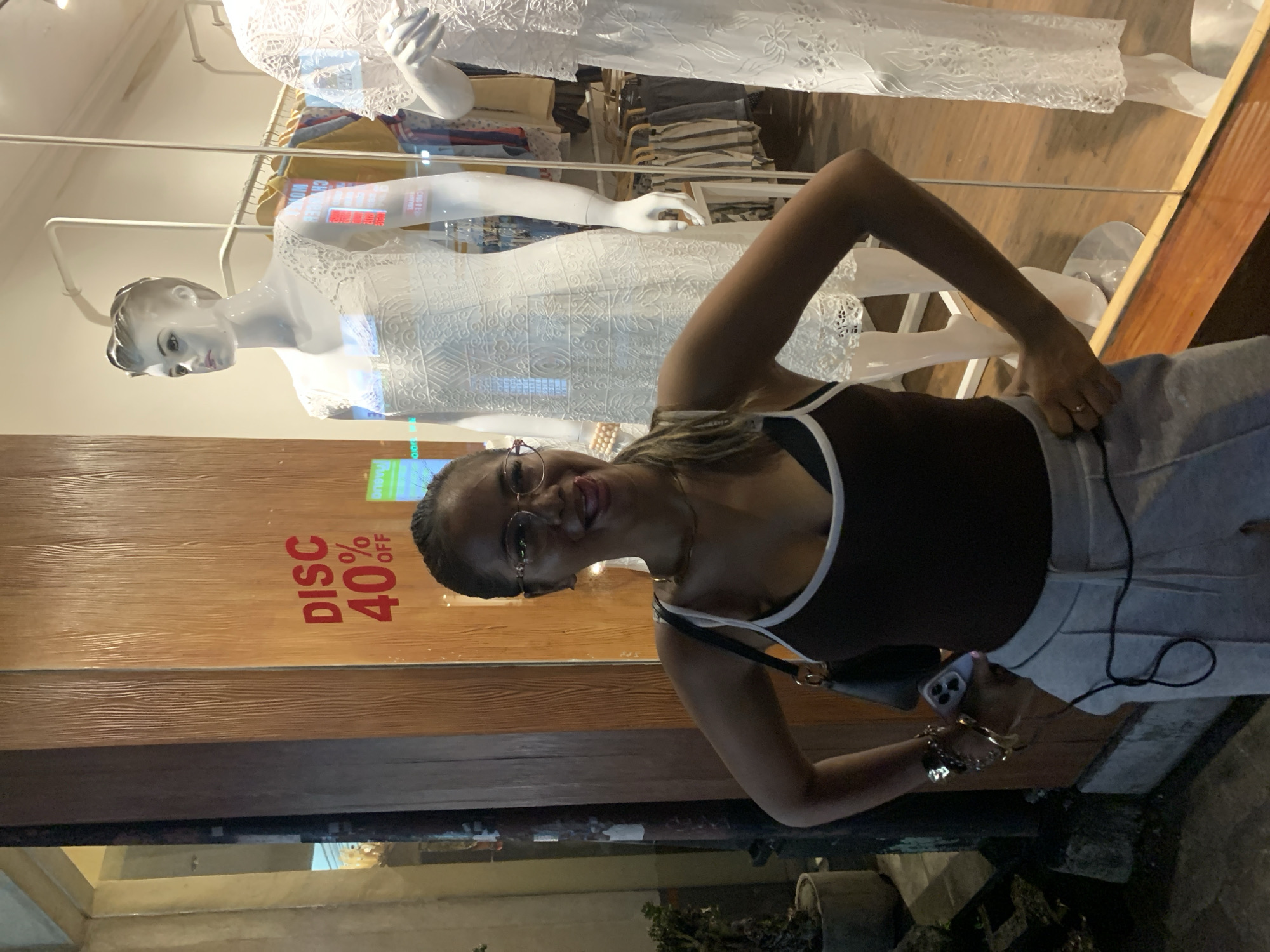 A woman is standing in front of a shop window with mannequins in Ubud District, Indonesia. The shop has a wooden panel with "DISC 40% OFF" printed on it. The woman is wearing glasses, a dark tank top with white trim, and grey pants. She is holding a phone in her left hand and has bracelets on her left wrist. Inside the shop window, there are several mannequins wearing white dresses. There are clothes racks with clothes hanging on them in the background. The environment outside the shop is dimly lit with some plants visible at the bottom of the frame.