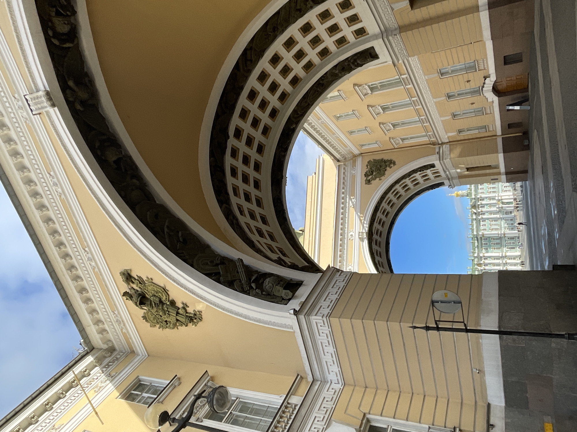 The image captures a view through an archway in Saint Petersburg, Russia, likely the General Staff Building's triumphal arch, leading to a view of the Winter Palace and Palace Square. The architecture is grand and neoclassical, with large arches and ornate facades painted in a pale yellow. Intricate decorative elements, including a double-headed eagle crest and classical friezes, are visible on the building's exterior. The archway itself is decorated with coffers, and a darker, more textured band runs along its curve.
In the foreground, on the left, a lamppost stands on a cobbled pavement. A circular sign is attached to the lamppost. The sky above is partly cloudy, with patches of blue. Through the arch, the expansive Palace Square is visible, with the magnificent Winter Palace as the dominant structure. The Winter Palace is a large, green and white palace with numerous windows and a golden dome. A few indistinct figures, presumably pedestrians, are scattered across the square, suggesting a public space. The lighting indicates it is daytime, with sunlight illuminating the yellow facades and casting some shadows. The overall scene conveys a sense of historical grandeur and architectural significance.