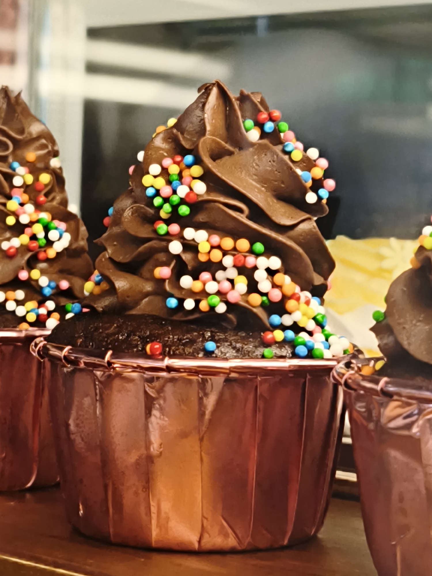This media file features a close-up, eye-level shot of three chocolate cupcakes with chocolate frosting and colorful sprinkles. The cupcakes are presented in rose gold wrappers, which are crimped at the top and pleated vertically. The frosting is swirled generously on top of each cupcake, and the sprinkles are scattered across the frosting and some have fallen onto the cake itself. The cupcakes appear to be on a wooden surface, and in the background, there's a blurred view of what looks like a display case with some yellow and white objects, possibly more baked goods. The lighting is bright, suggesting an indoor setting, possibly a bakery or a confectionery shop. The focus is sharp on the cupcakes, drawing attention to their texture and vibrant toppings. There is no visible text in the image.