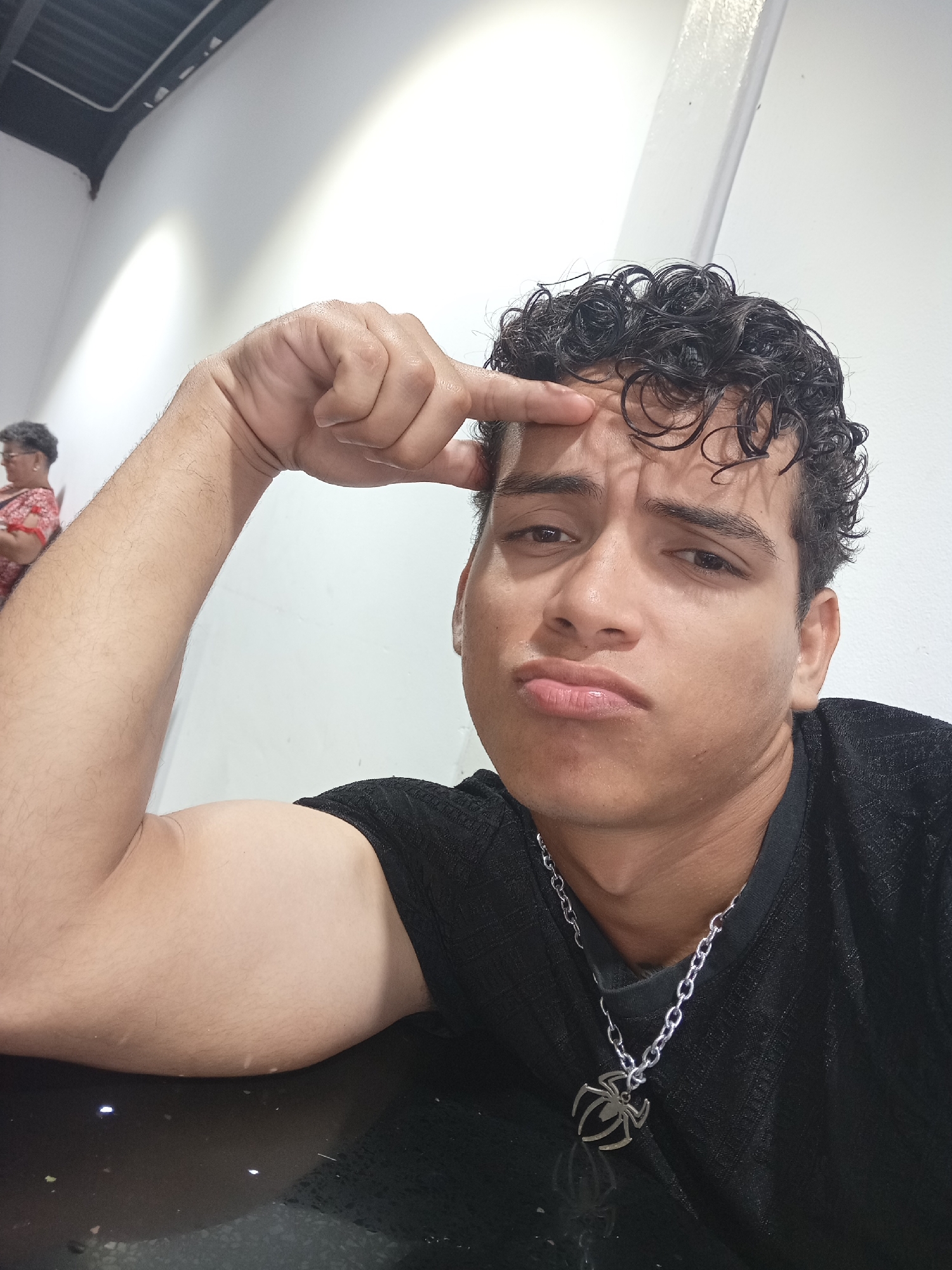 A young man with dark, curly hair and light brown skin is the central subject of this close-up, chest-up portrait. He appears to be in his late teens or early twenties and is wearing a black t-shirt and a silver chain necklace with a distinctive spider-shaped pendant. He is posing with his left arm resting on a dark, glossy surface, possibly a table, and his bicep is noticeably flexed. His right hand is raised, with his index finger touching his forehead, creating a furrowed brow and a pouting expression on his lips, suggesting a look of playful confusion, contemplation, or mild annoyance.
The setting is an indoor space, characterized by plain white walls and a dark ceiling with visible lighting fixtures, suggesting a modern or minimalist environment. In the softly blurred background, an older woman with short, grey hair, wearing a red and white patterned top, is partially visible, indicating a public or semi-public space like a restaurant or cafeteria. The lighting is bright and even, likely artificial, with no strong shadows or natural light cues to indicate the time of day beyond a well-lit interior. No visible text is present in the image. The overall scene captures a casual, posed moment, likely taken in Juan Griego, Venezuela.