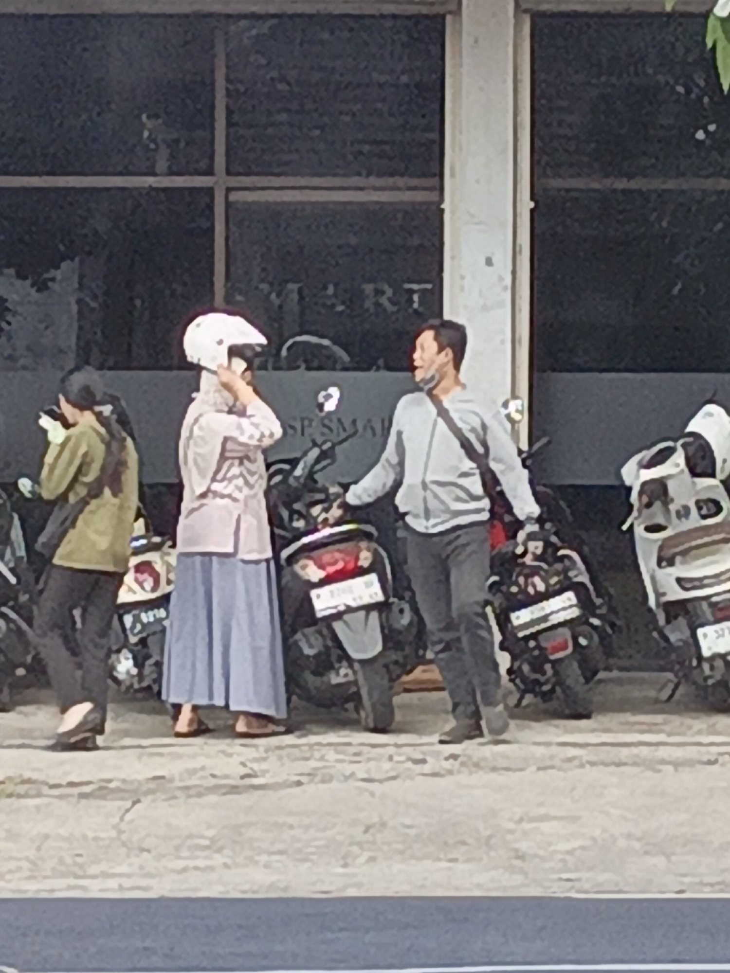In Srono, Indonesia, on a sunny day, two individuals are interacting near a row of parked motorcycles outside a building. A person wearing a white helmet, a light-colored headscarf, a patterned top, and a blue skirt is adjusting their helmet. To their right, a man in a grey hooded jacket and dark pants, with a dark bag slung across his chest, is gesturing with his left hand while looking to his left. A third person, dressed in a green jacket and dark pants, stands to the far left, facing away from the camera, holding a light-colored object. The ground is paved and cracked, with a dark blue stripe running along the bottom edge of the frame. The background features dark tinted windows of a building.
