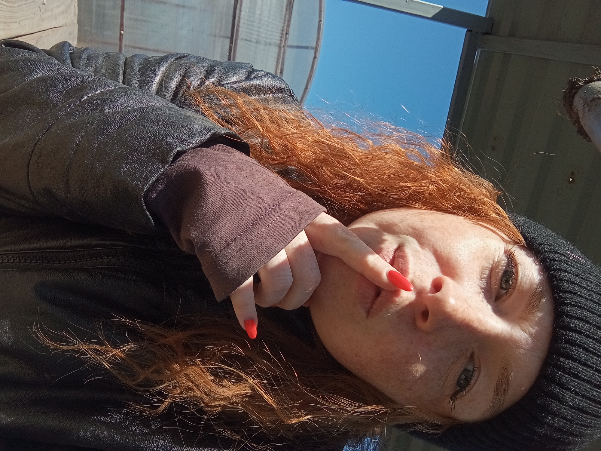 A woman with red hair and fair skin lies on her side, facing the camera. She wears a black knit beanie and a black leather jacket with a brown sleeve visible underneath. Her fingernails are painted bright orange-red. She holds one finger close to her lips. The background shows a blue sky and parts of a metallic structure with transparent panels, suggesting a greenhouse or covered area. The location is possibly in Yurgamysh, Russia.