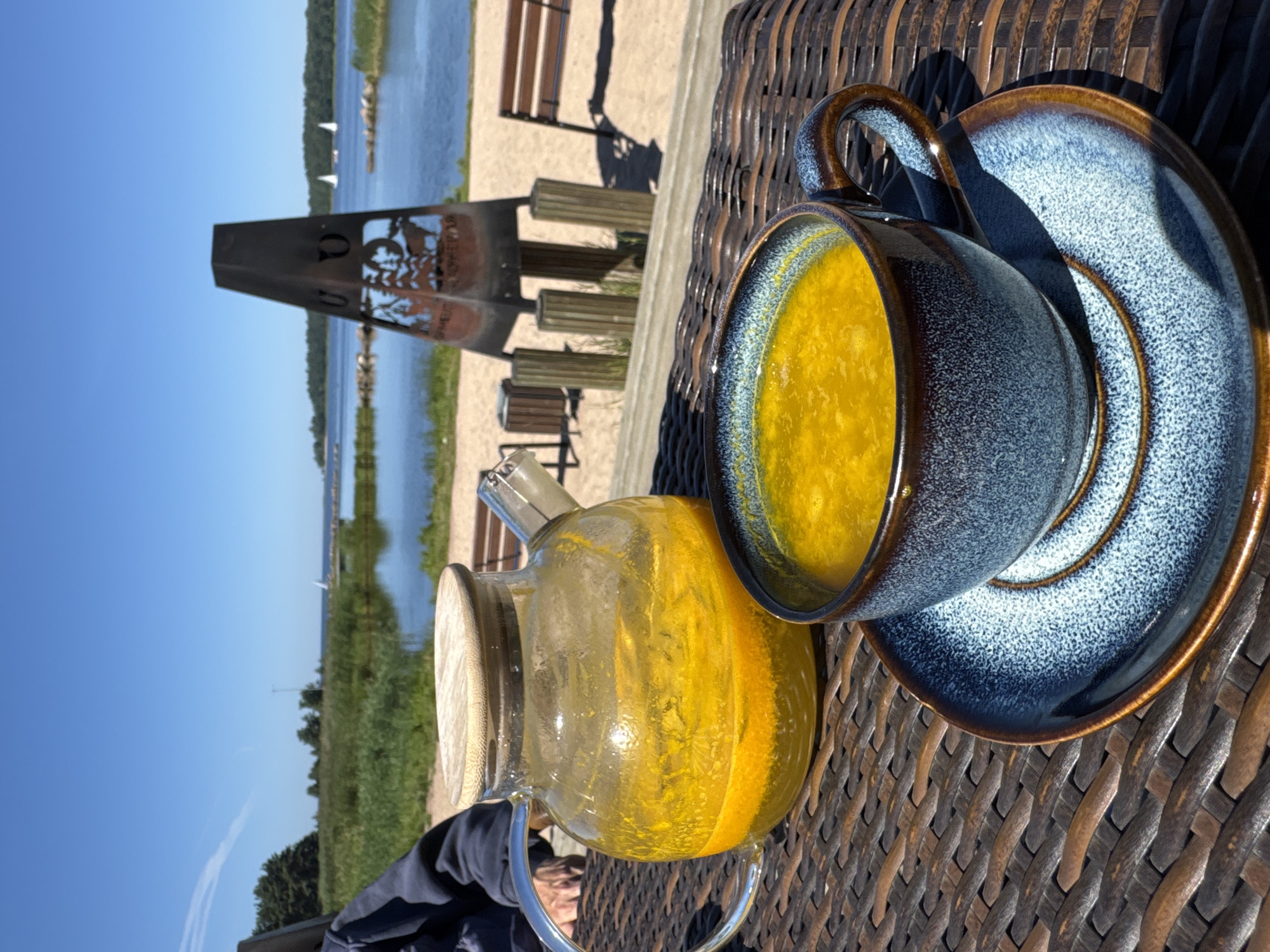 An eye-level, medium shot captures a table setting by a body of water, likely a lake or sea, on a sunny day. In the foreground, a woven brown table holds a clear glass teapot filled with a bright yellow liquid, possibly a beverage or infused water. Next to it, a blue ceramic mug with a brown rim, also filled with the yellow liquid, sits on a matching saucer. The background reveals a sandy shore with some vegetation and a metallic sculpture of a pyramidal shape featuring cut-out silhouettes of animals and trees. Beyond the shore, a calm blue body of water stretches out, dotted with several white sailboats. The sky is a clear, bright blue, with a faint white cloud streak. The setting suggests an outdoor cafe or a recreational area, possibly in Ozerki (Krasnodolinskaya), Russia, given the context provided. A person's arm, wearing a dark jacket, is visible on the left side of the frame, suggesting their presence at the table.