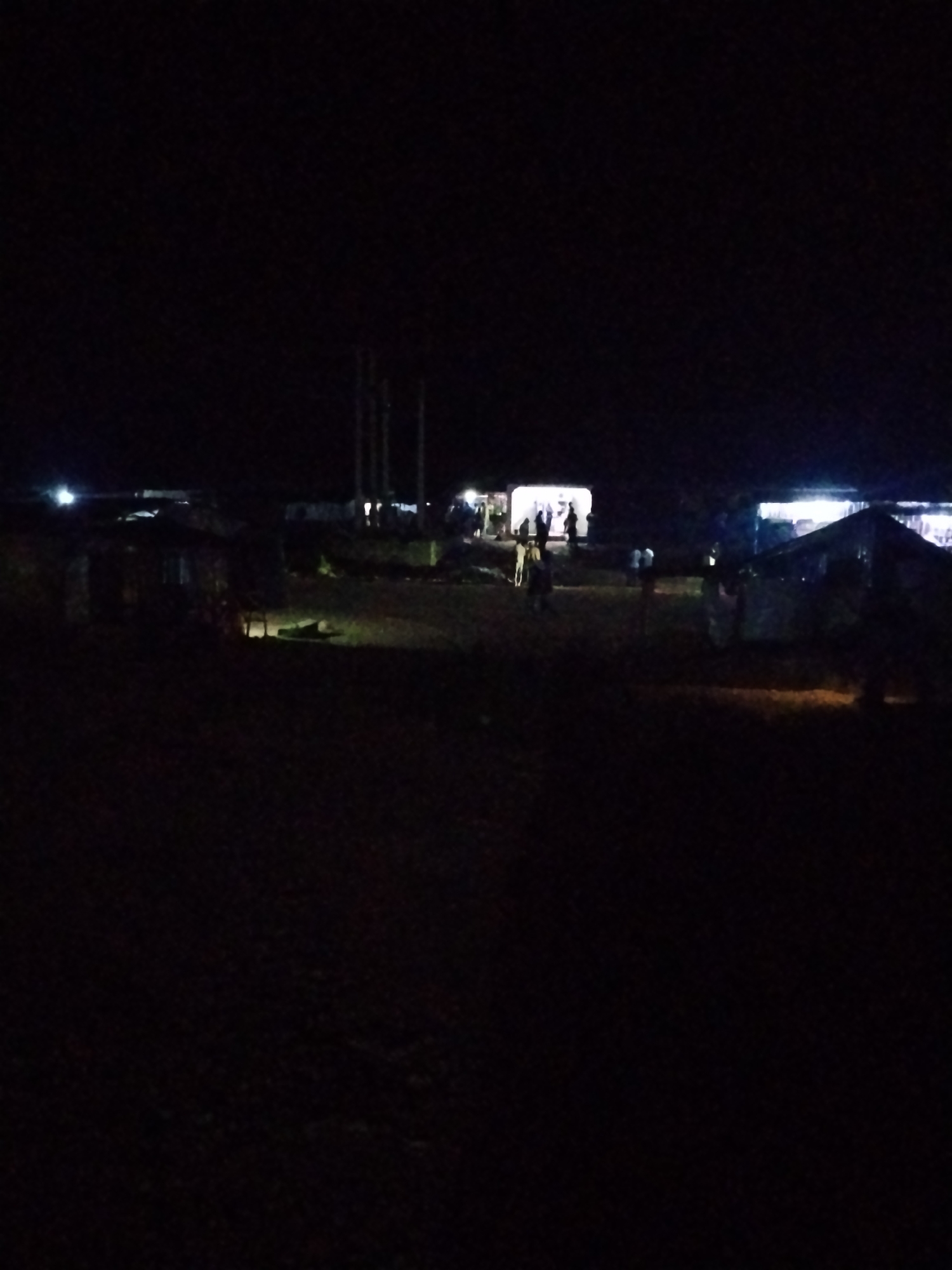 The image depicts a nighttime scene in a rural or informal settlement in Durumi, Nigeria. Several structures, likely temporary or makeshift dwellings, are visible, illuminated by artificial lights. A few individuals are present, some gathered around one of the illuminated buildings. The overall environment appears to be unpaved and dark, with the lights from the structures creating pools of illumination.