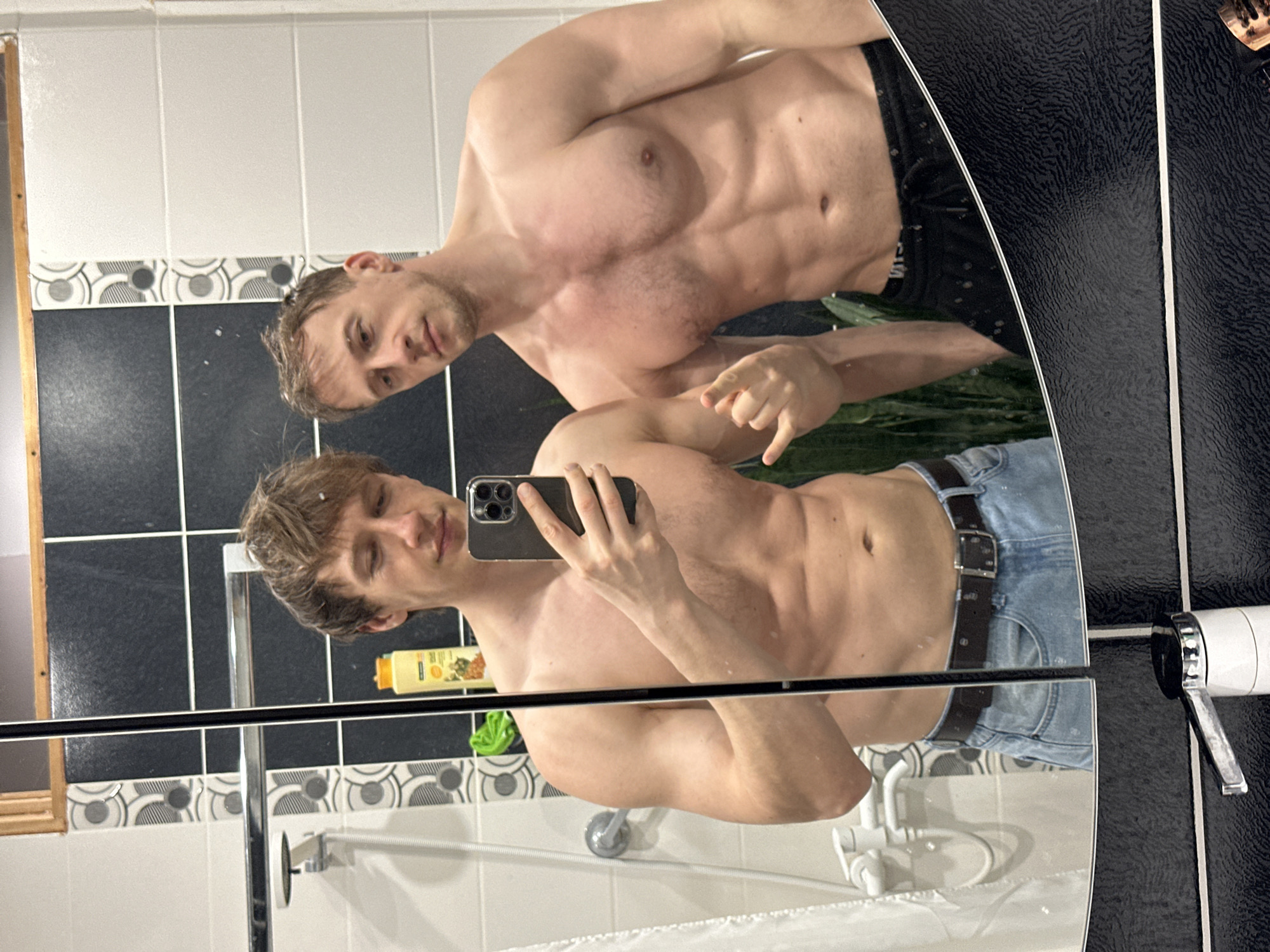 A shirtless man with blond hair and light complexion is taking a mirror selfie in a bathroom. The bathroom has black and white tiled walls and a shower. The reflection in the mirror shows another man without a shirt, indicating they are together. The location is likely a private bathroom. The city of Kursk, Russia, is not verifiable from the image.