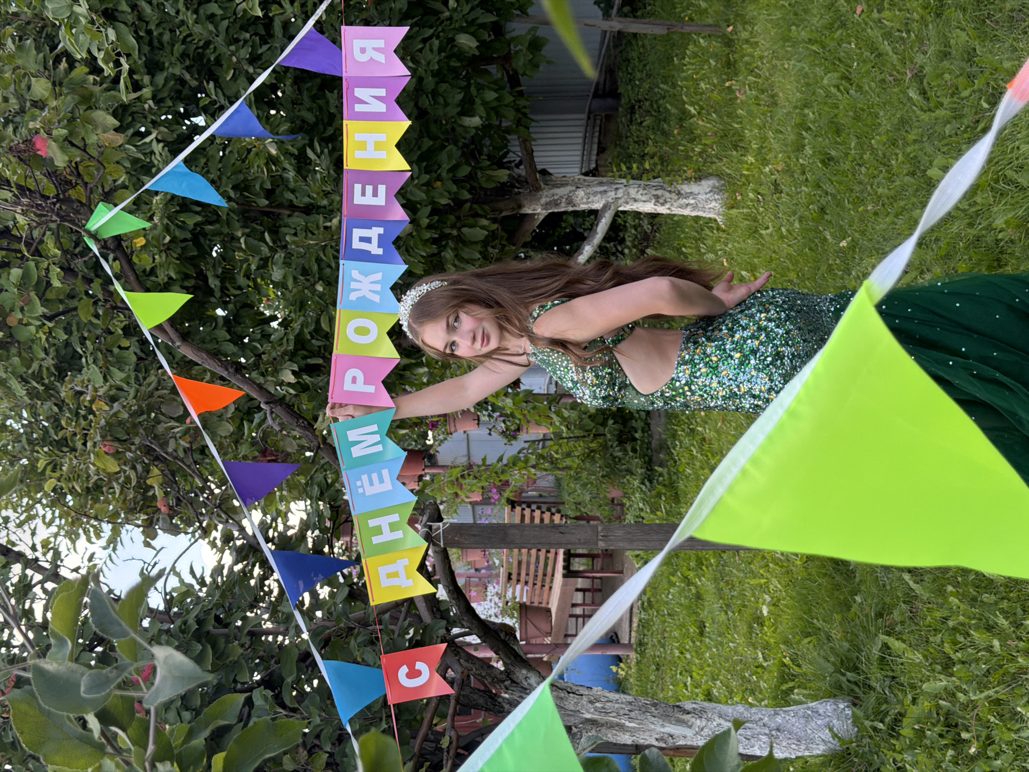 The image depicts a young woman with long, brown hair, wearing a sparkly green dress and a tiara, standing in a grassy outdoor area. She is posing with her arm raised, holding a banner that reads "С ДНЁМ РОЖДЕНИЯ" (Happy Birthday in Russian). The banner is strung between trees and decorated with colorful triangular flags. In the background, there are trees, a wooden bench, potted plants, and a fence. The scene suggests a birthday celebration or party taking place in a backyard or garden. There is no specific indication of the location within the city of Chaikovsky, Russia, beyond it being an outdoor setting.