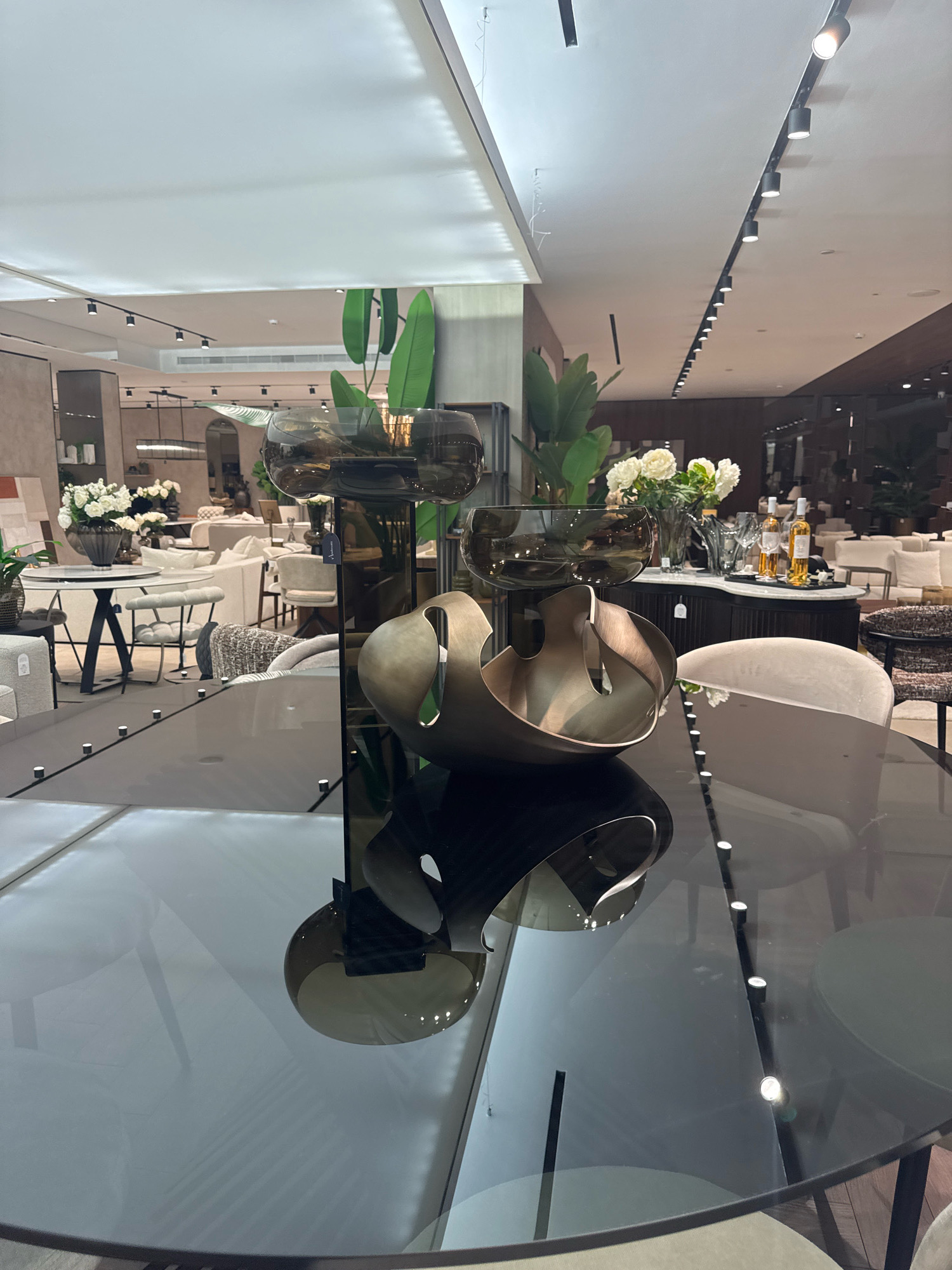 The image depicts a high-end furniture showroom in Agios Athanasios Municipality, Cyprus. In the foreground, a sleek, dark glass dining table with embedded lights dominates the frame. Reflecting on its surface are two abstract, metallic sculptures, one appearing to be a large, open bowl shape with swirling elements, and another, a darker, more solid form. Behind the table, a diverse range of modern furniture is displayed, including plush sofas, armchairs upholstered in neutral fabrics, and round dining tables with matching chairs. Several arrangements of white flowers in vases add a touch of elegance to the scene. In the background, track lighting illuminates the ceiling, and large windows offer glimpses of other sections of the showroom, showcasing more furniture and architectural elements. The overall impression is one of luxury and sophisticated design.
