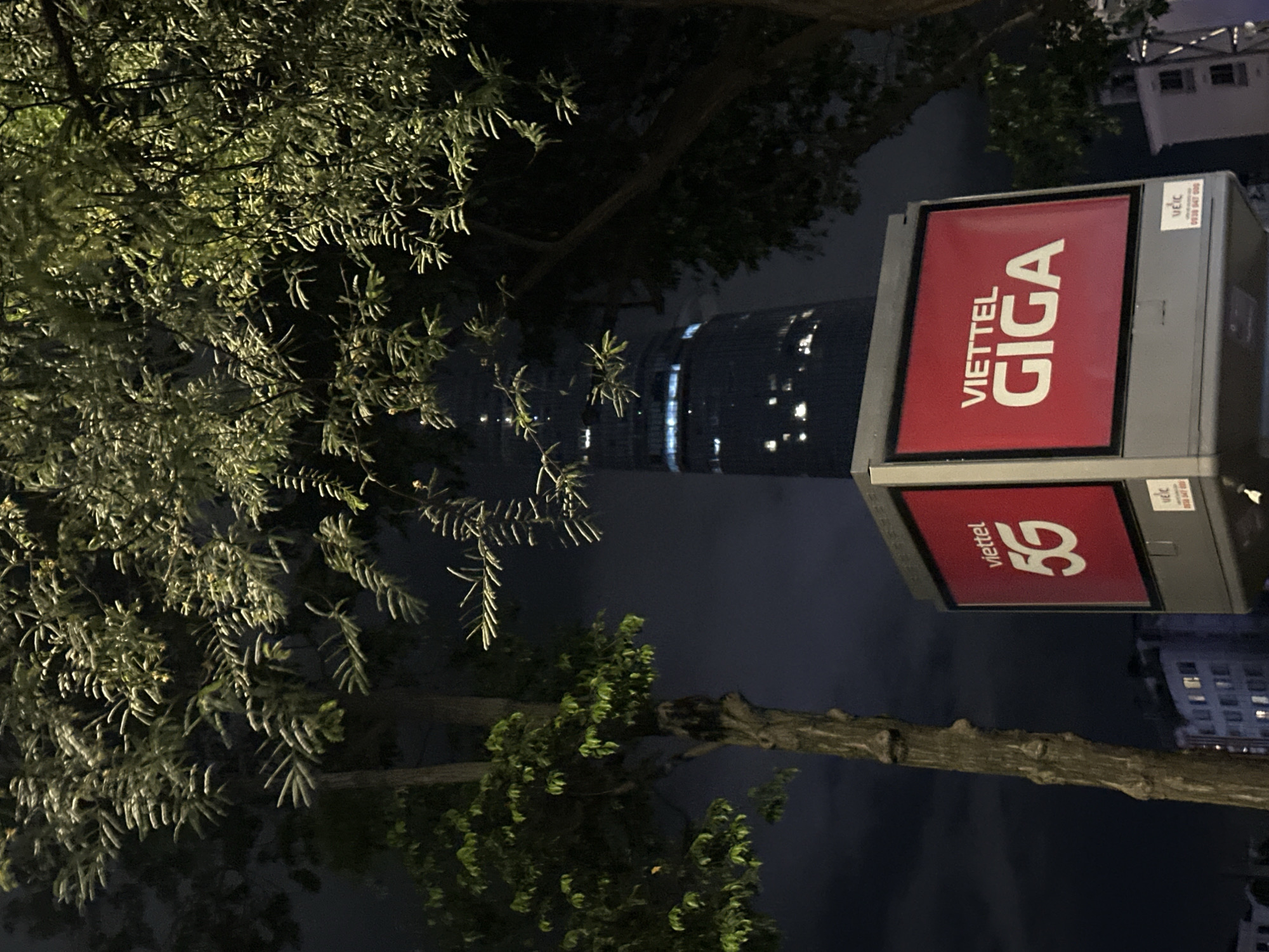 At night in Thủ Đức, Vietnam, a tall, dark skyscraper with illuminated windows rises in the background, partially obscured by the branches of a lush green tree. In the foreground, a digital billboard with a red background displays white text. The left side of the billboard shows "Viettel" above "5G". The right side of the billboard reads "VIETTEL" above "GIGA". Below the right side of the billboard, a smaller white sticker displays some text, including "0938 947 999". The scene is illuminated by artificial lights, casting a cool glow on the foliage and the billboard.
