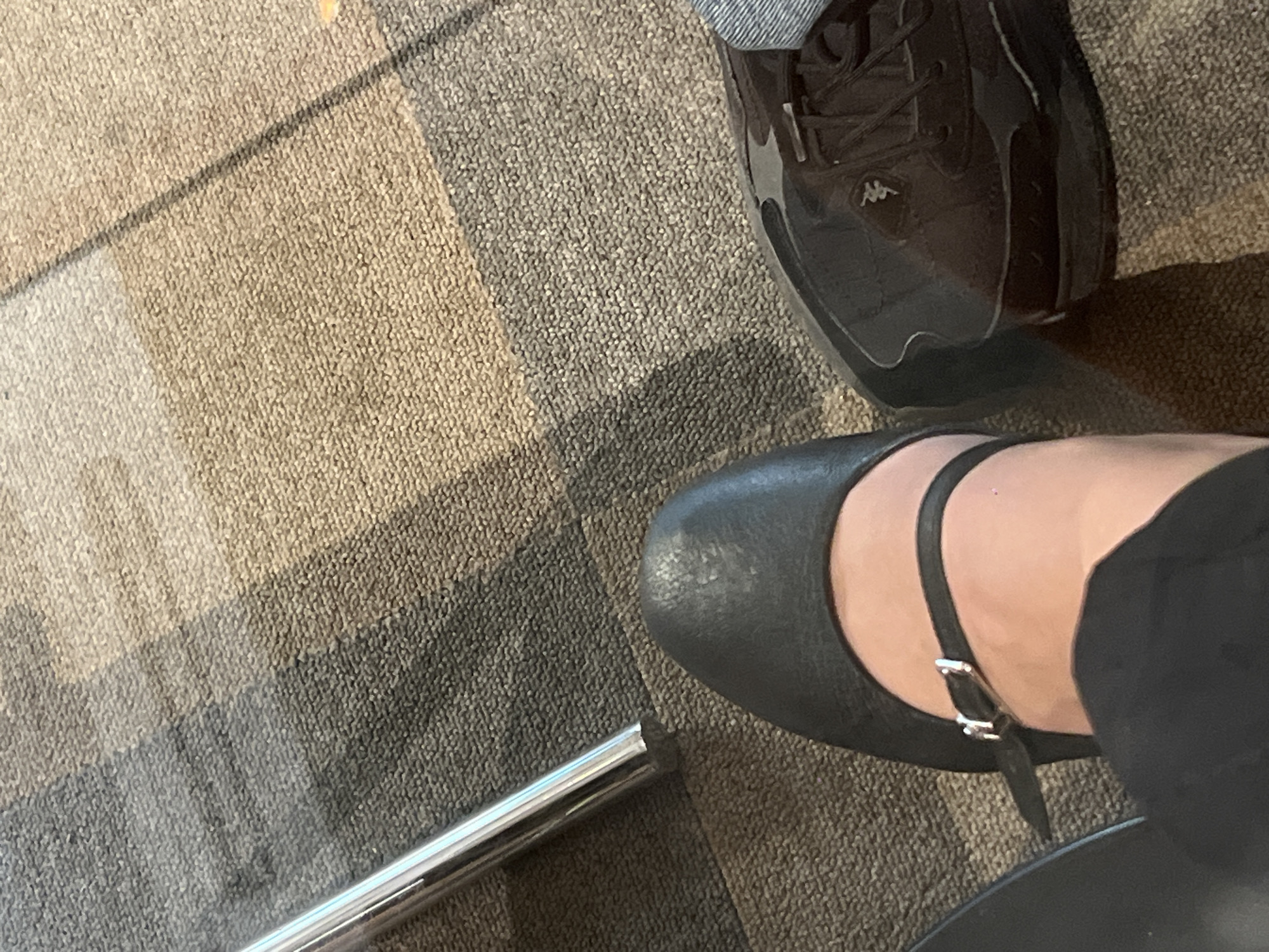 The image shows a close-up view of two pairs of shoes on a carpeted floor. One pair is black athletic shoes, and the other is black leather Mary Jane-style shoes with an ankle strap. A metallic object, possibly part of a chair or furniture, is visible near the Mary Jane shoe. There are no people visible other than the feet/lower legs of the individuals wearing the shoes. The location is not discernible.
